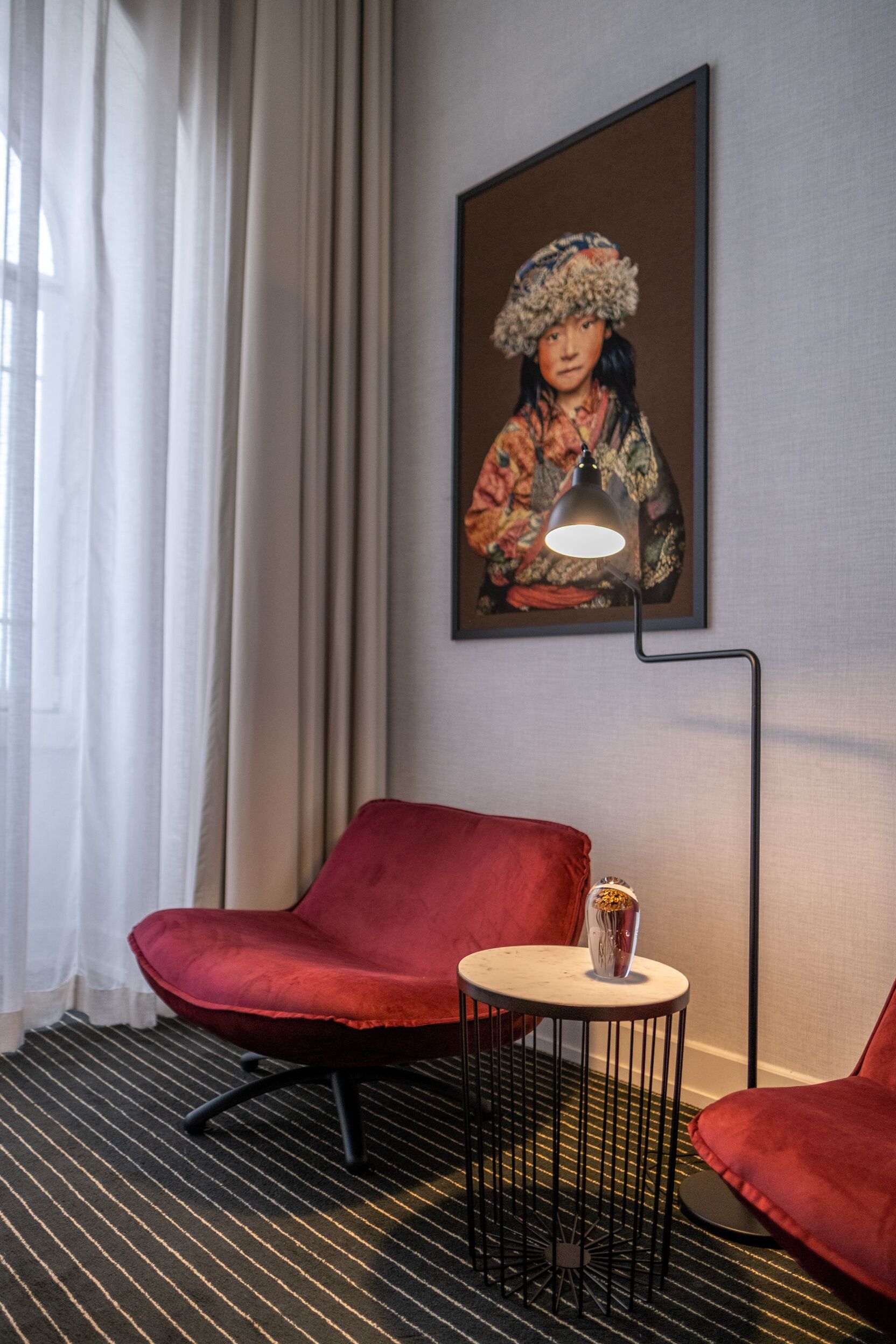 A hotel room with a red chair, a small round table with a vase, and a painting on the wall.