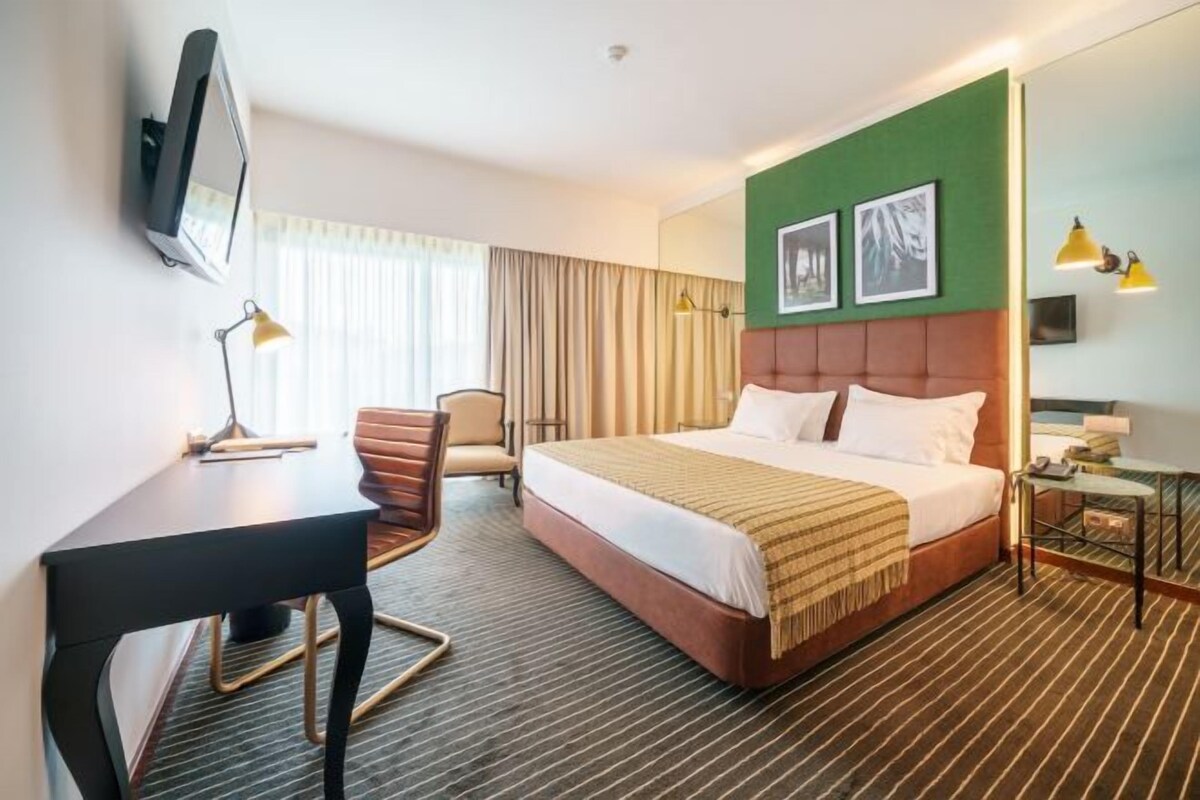 A hotel room with a bed, a desk, a chair, a TV, and a small table with a lamp.