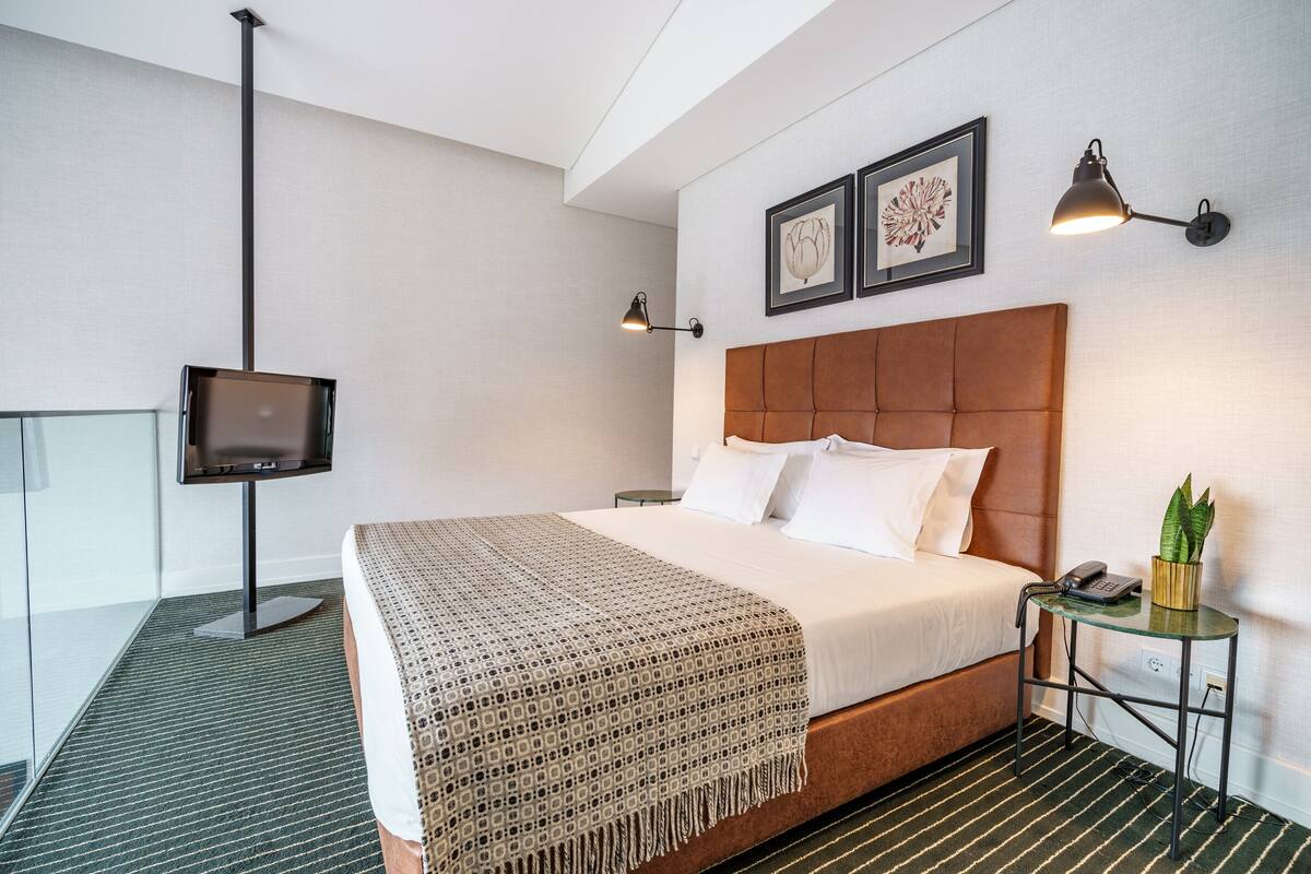 A modern hotel room with a large bed, a flat-screen TV, two wall-mounted lamps, and a small side table with a plant.