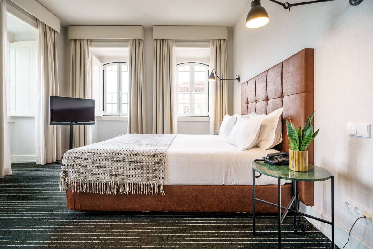 A hotel room with a large bed, a bedside table with a plant, a flat-screen TV, and a window with curtains.