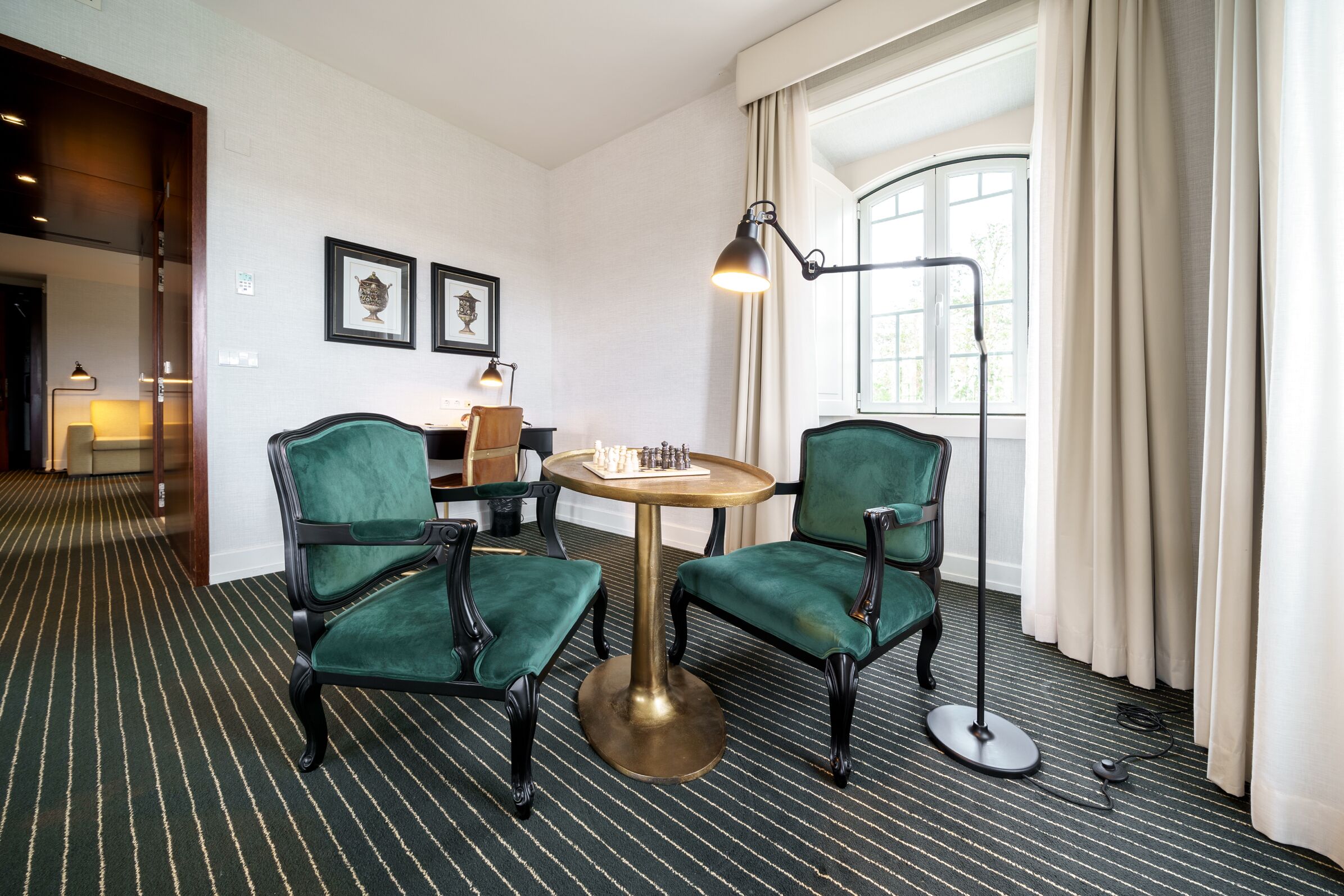 Habitación de hotel con una butaca tapizada de verde, una mesa redonda, una lámpara de pie y una ventana con cortinas.