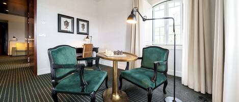 Habitación de hotel con una butaca tapizada de verde, una mesa redonda, una lámpara de pie y una ventana con cortinas.