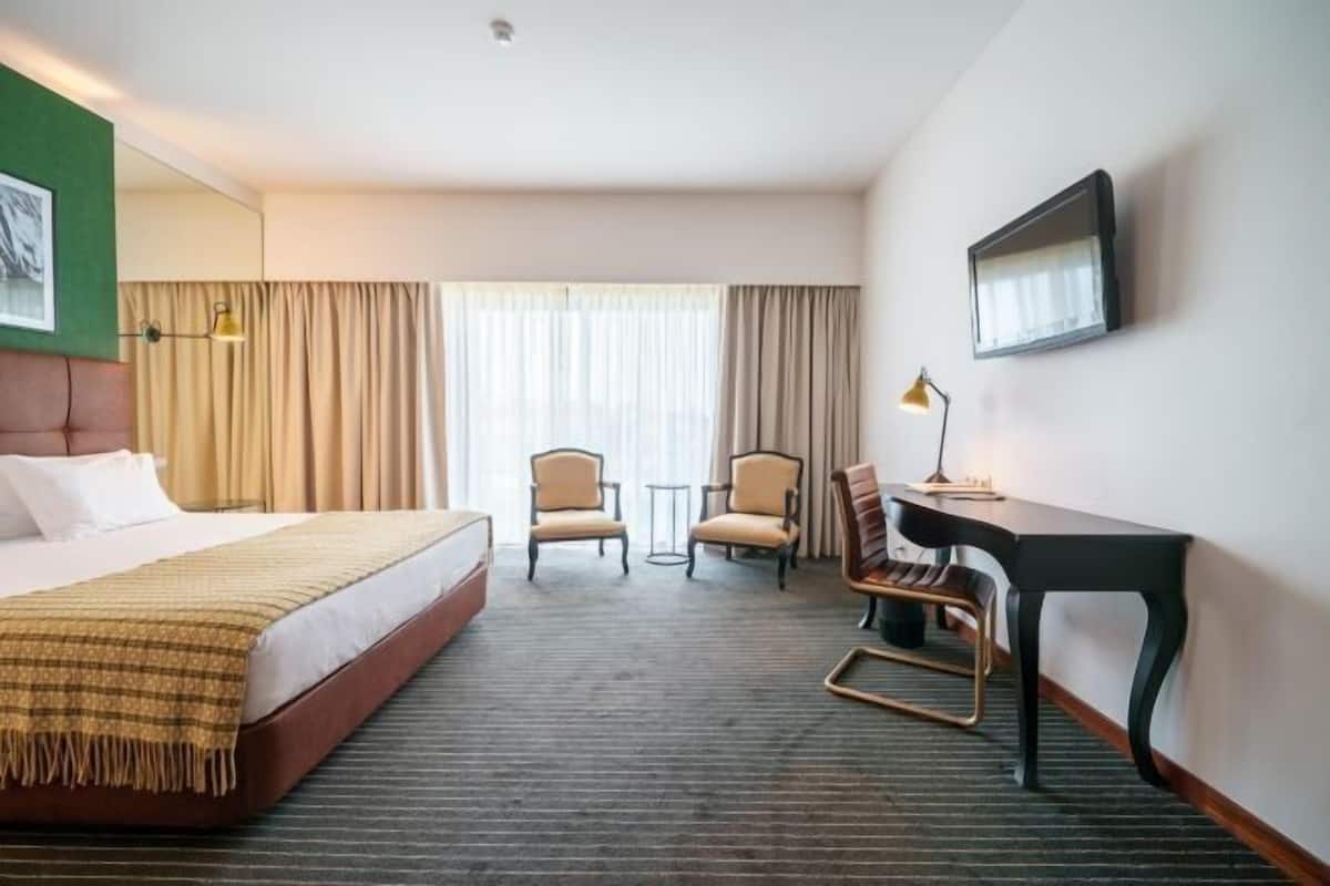 A hotel room with a bed, two chairs, a desk, a television, and a window with curtains.