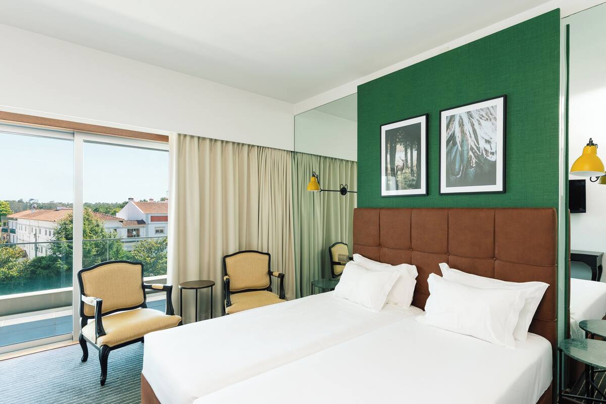 A hotel room with a large bed, two chairs, a small table, a mirror, and a view of the city.