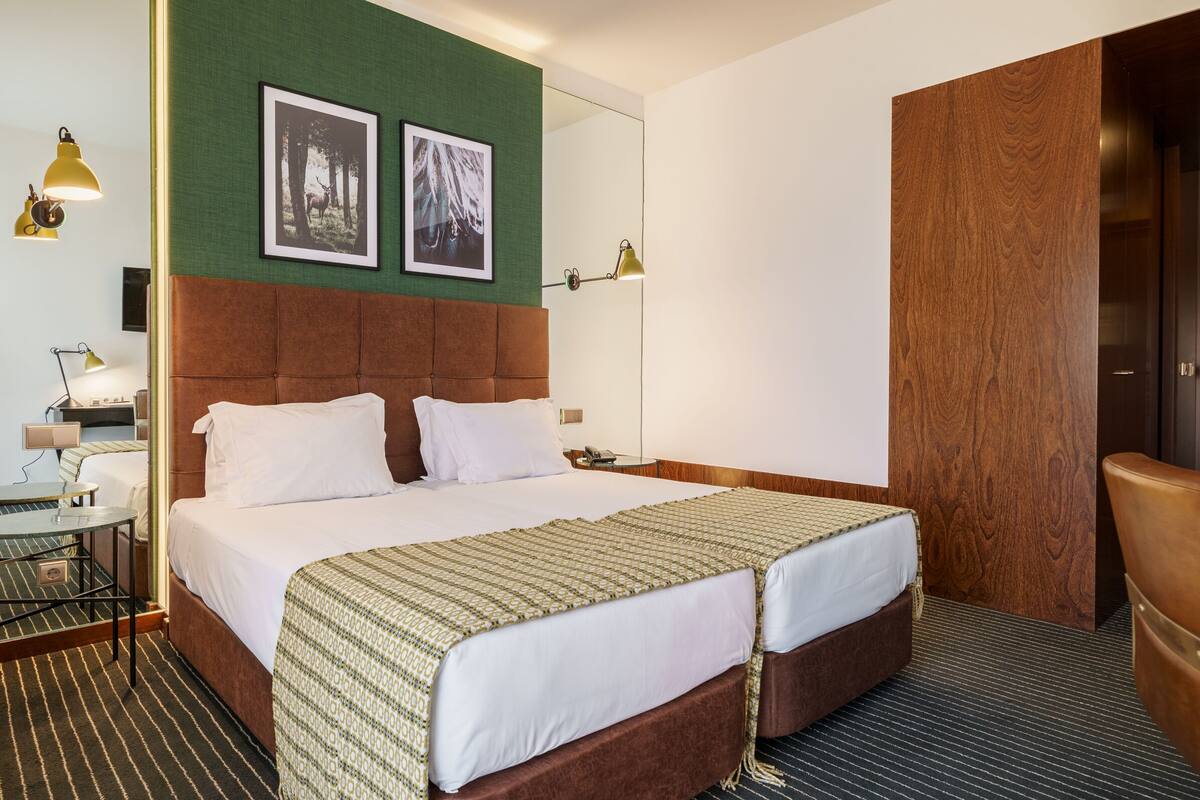 A hotel room with a large bed, two bedside tables, a desk, and a chair. There are two framed pictures on the wall, a lamp, and a wooden panel on the door.
