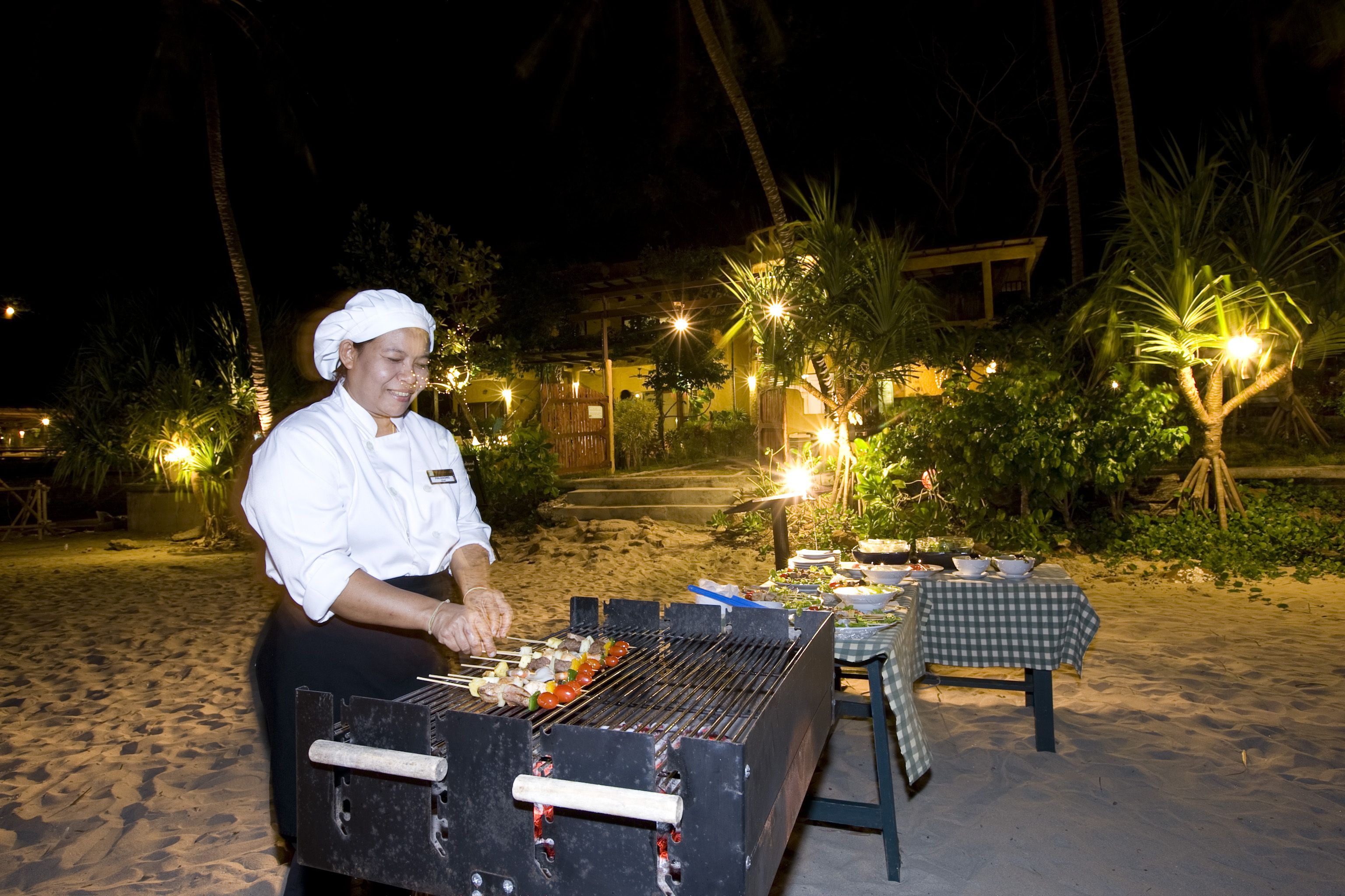 A chef grilling outdoors at night with a table set for dining nearby.