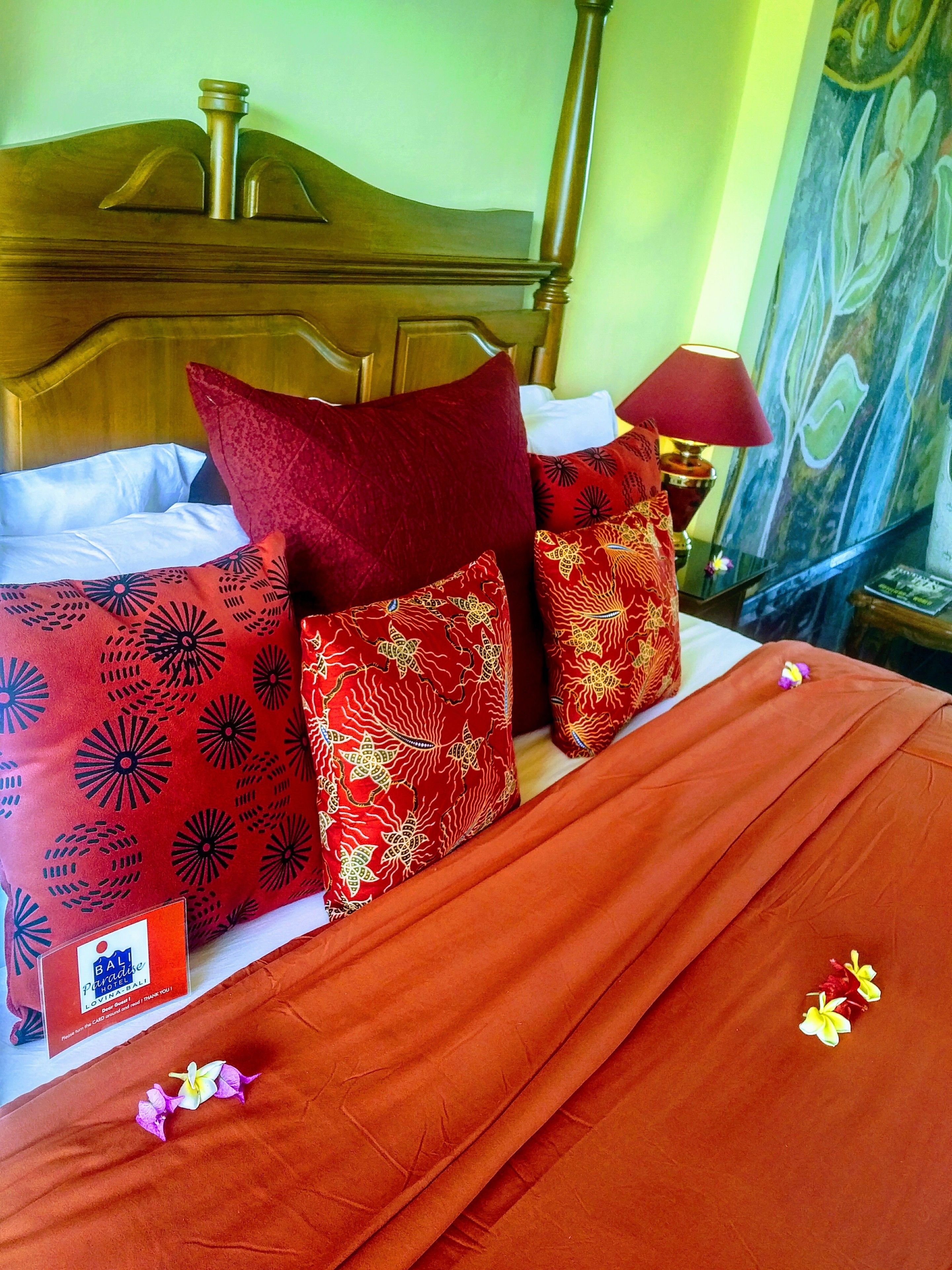 Habitación de hotel con cabecera de madera, almohadas estampadas rojas, manta roja y mesita de noche con lámpara.