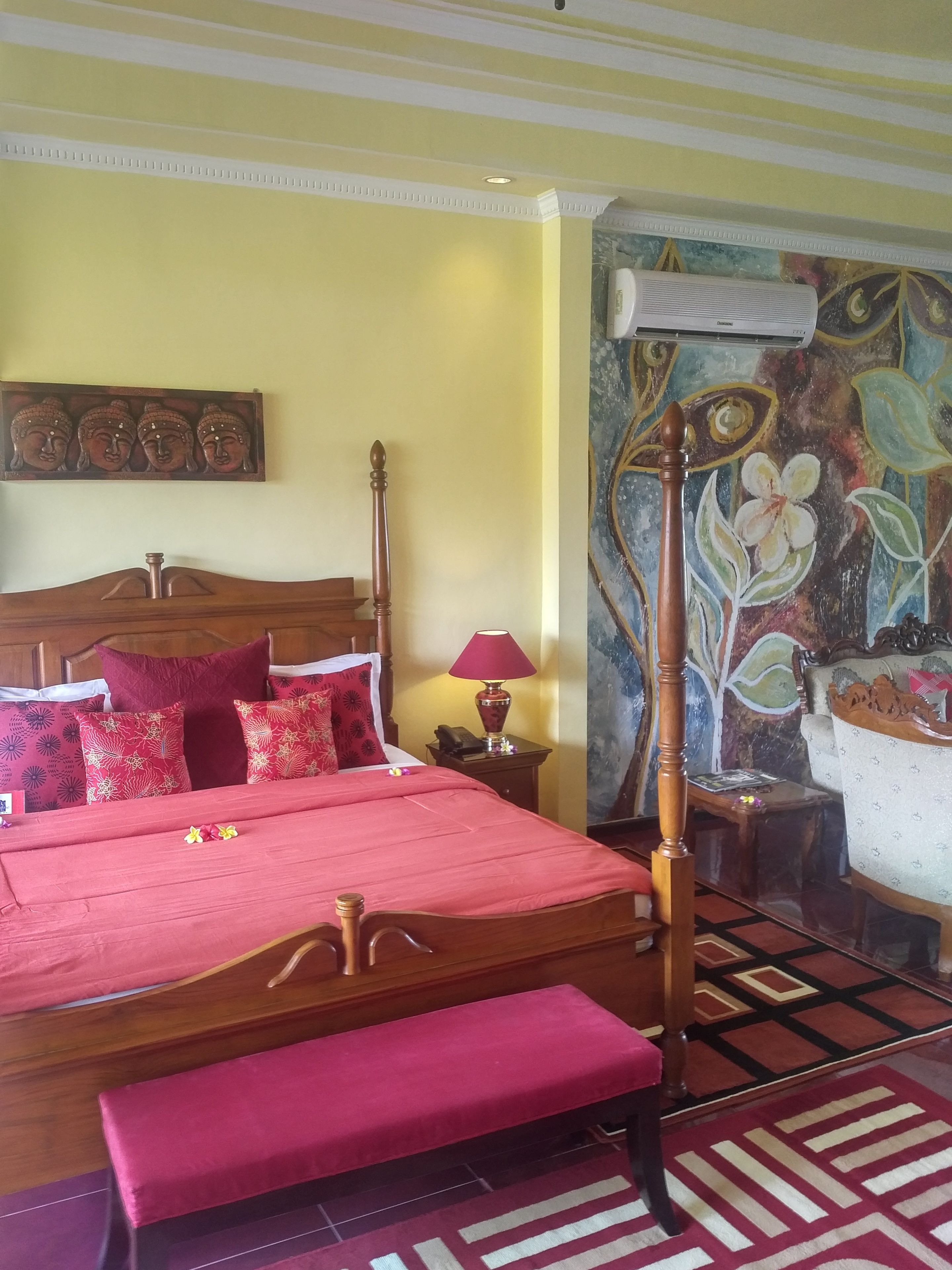 Une chambre d’hôtel avec un lit en bois, un banc, une table de chevet avec une lampe et un mur orné d’une fresque florale.