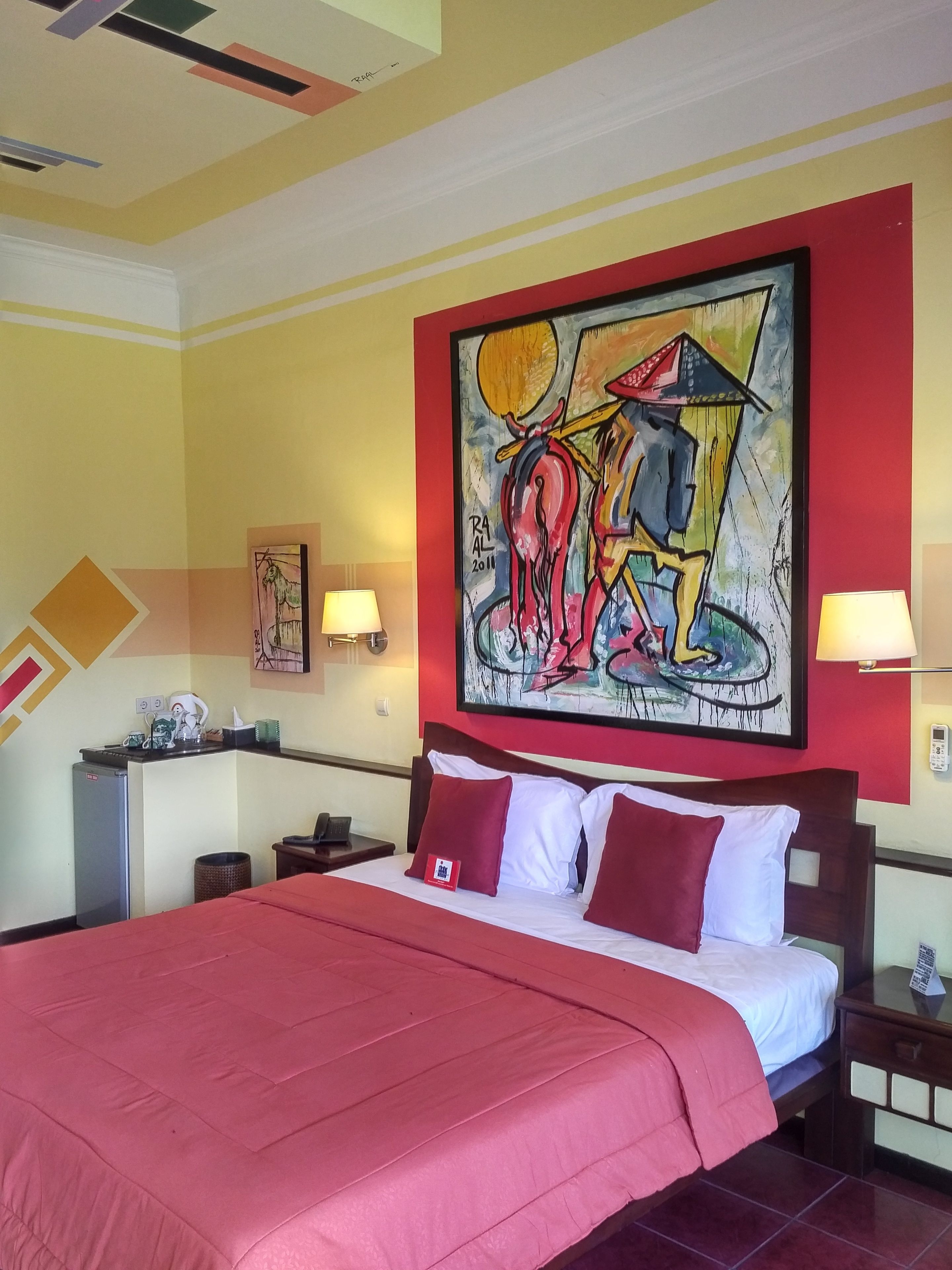 Habitación de hotel con una cama grande, un escritorio con lámpara, un refrigerador y un cuadro en la pared.