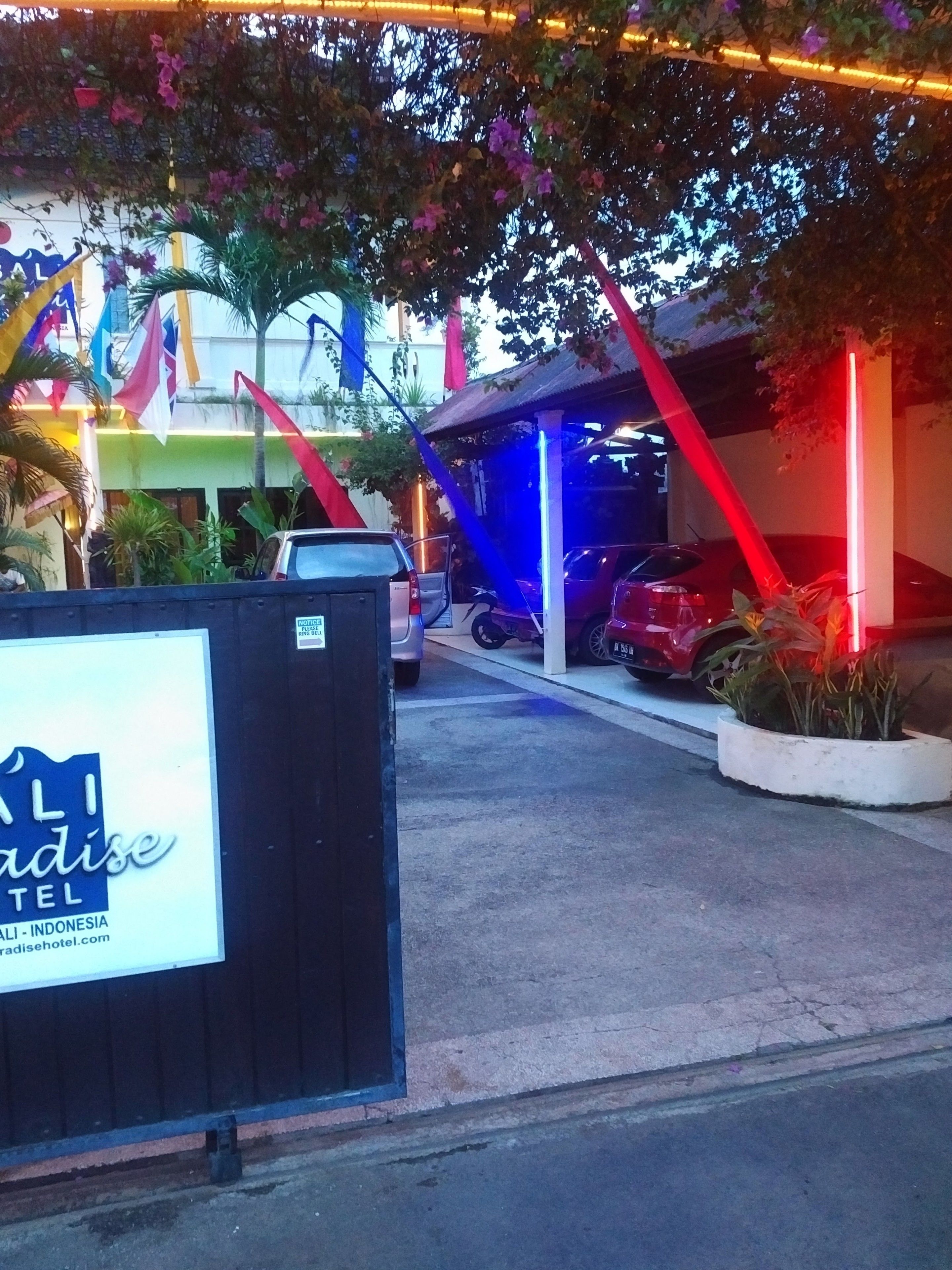 A hotel entrance with a signboard, parked cars, and colorful decorations.