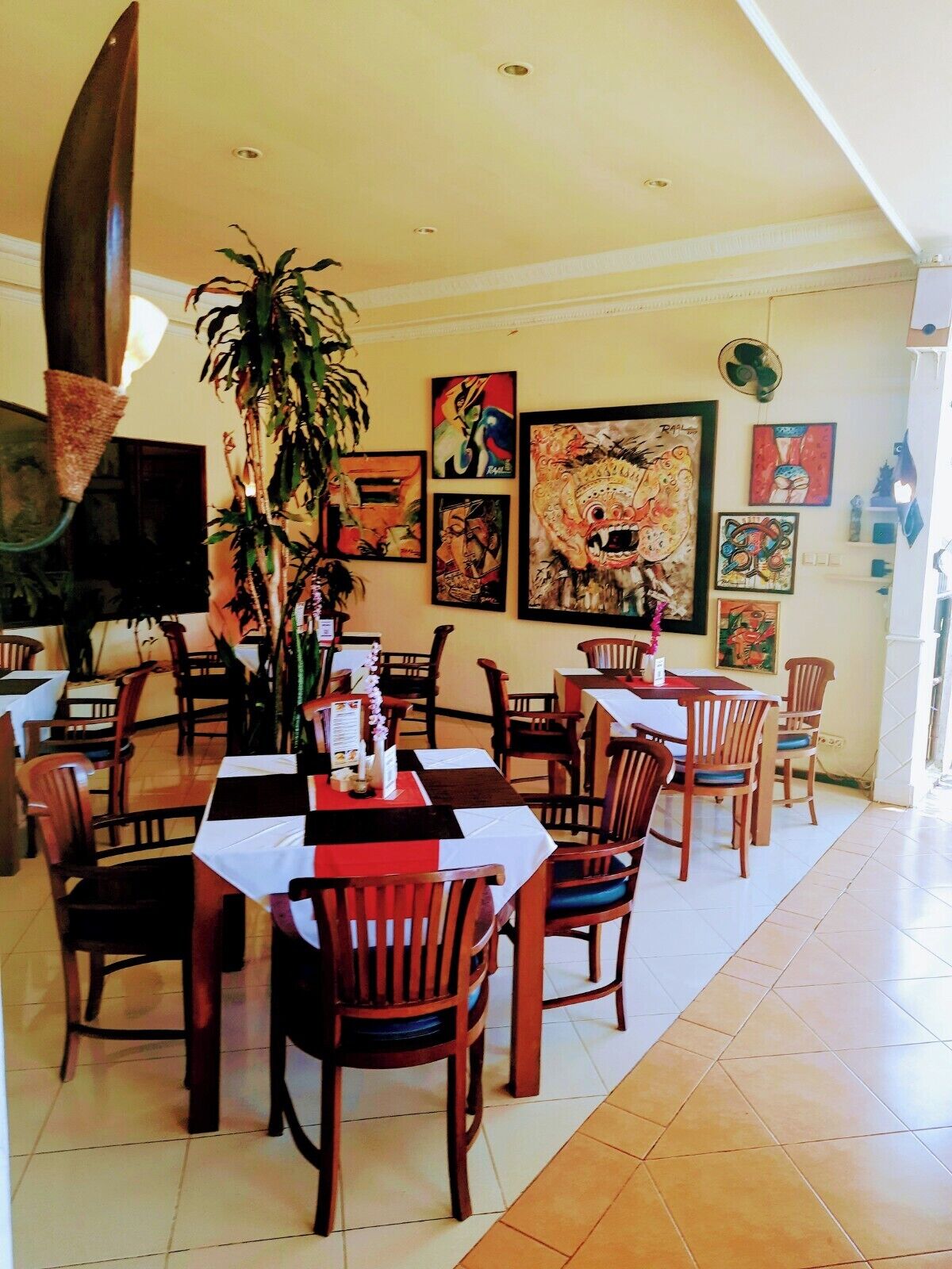 Un comedor con mesas y sillas de madera, una planta grande y varias obras de arte en las paredes.