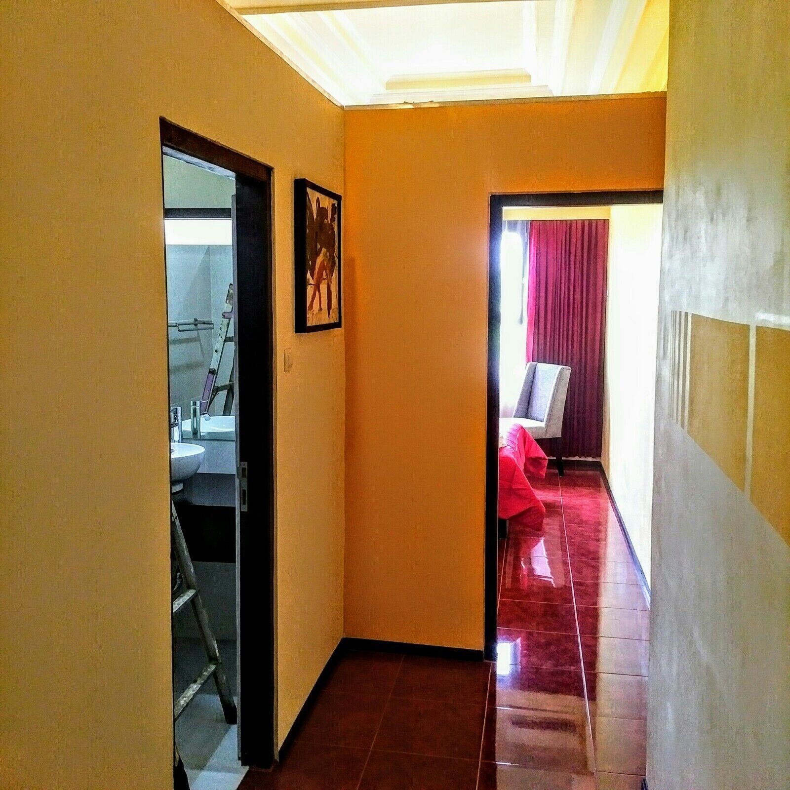 A hallway with a tiled floor, a door leading to a bathroom, and a view into a room with a bed and a window with curtains.