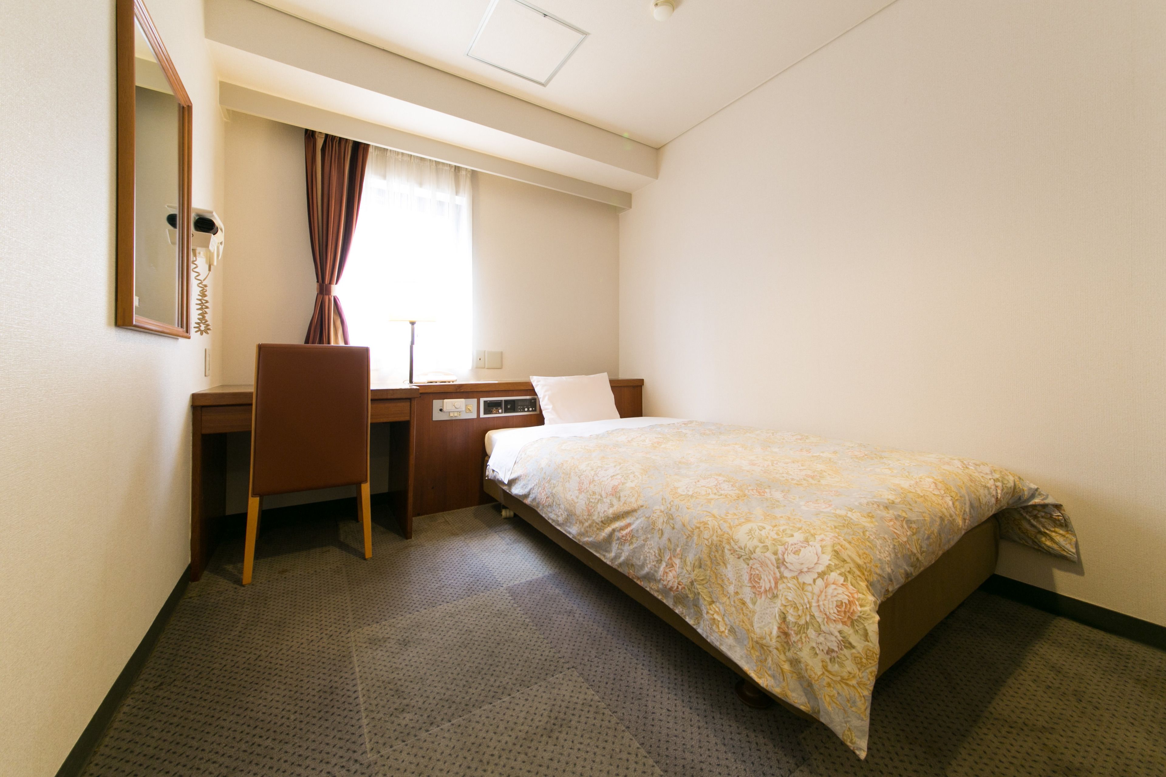A hotel room with a bed, a desk, a chair, a mirror, and a window with curtains.