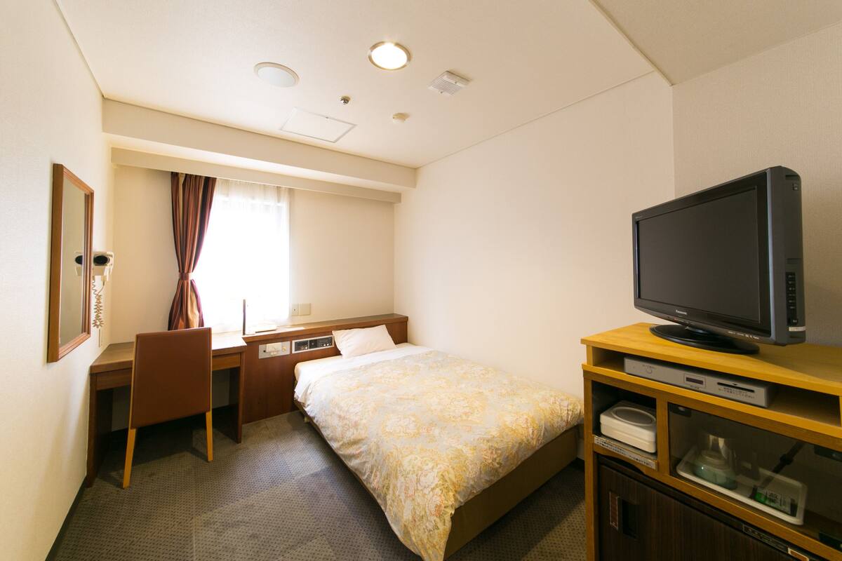 A hotel room with a bed, a desk with a chair, a television, and a minibar.