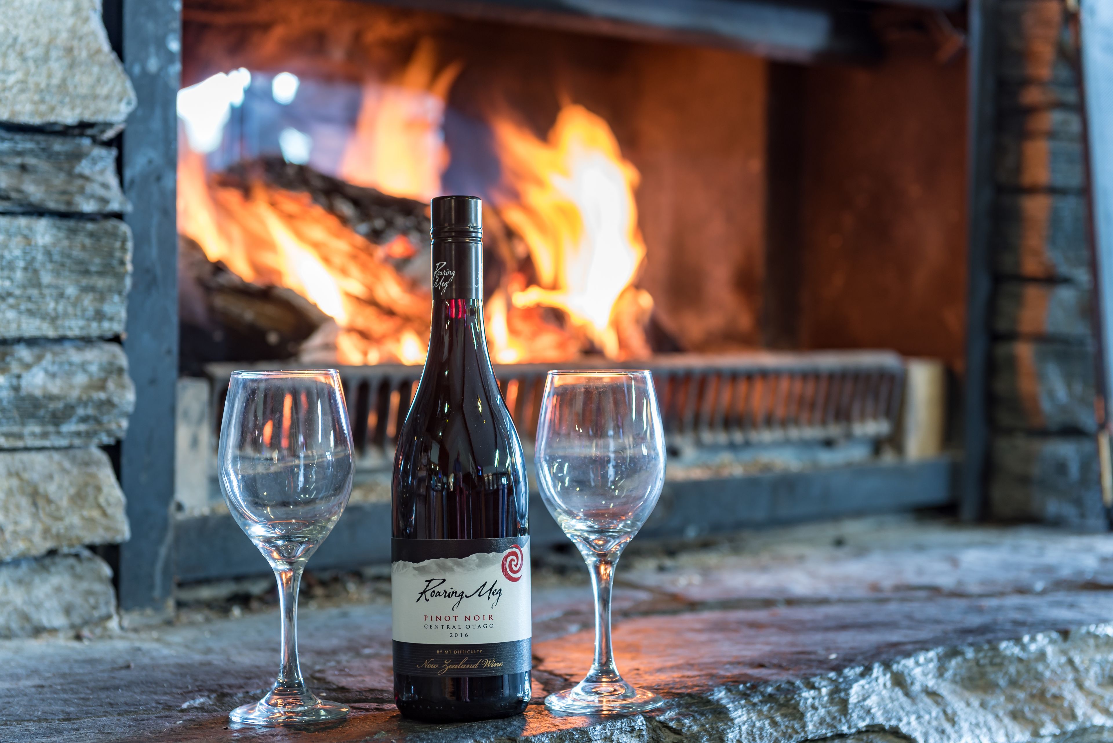 A bottle of wine and two glasses on a stone surface with a fireplace in the background.