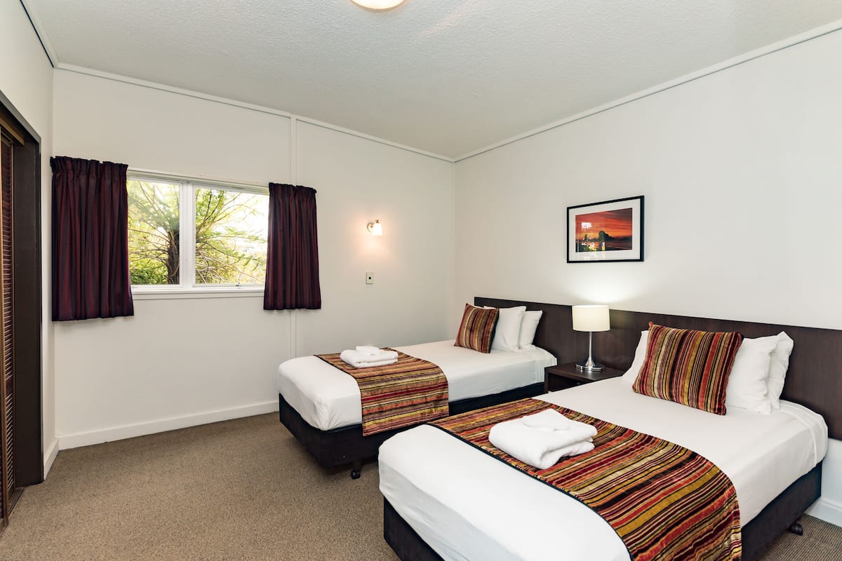 A hotel room with two beds, a window with curtains, a nightstand with a lamp, and a framed picture on the wall.