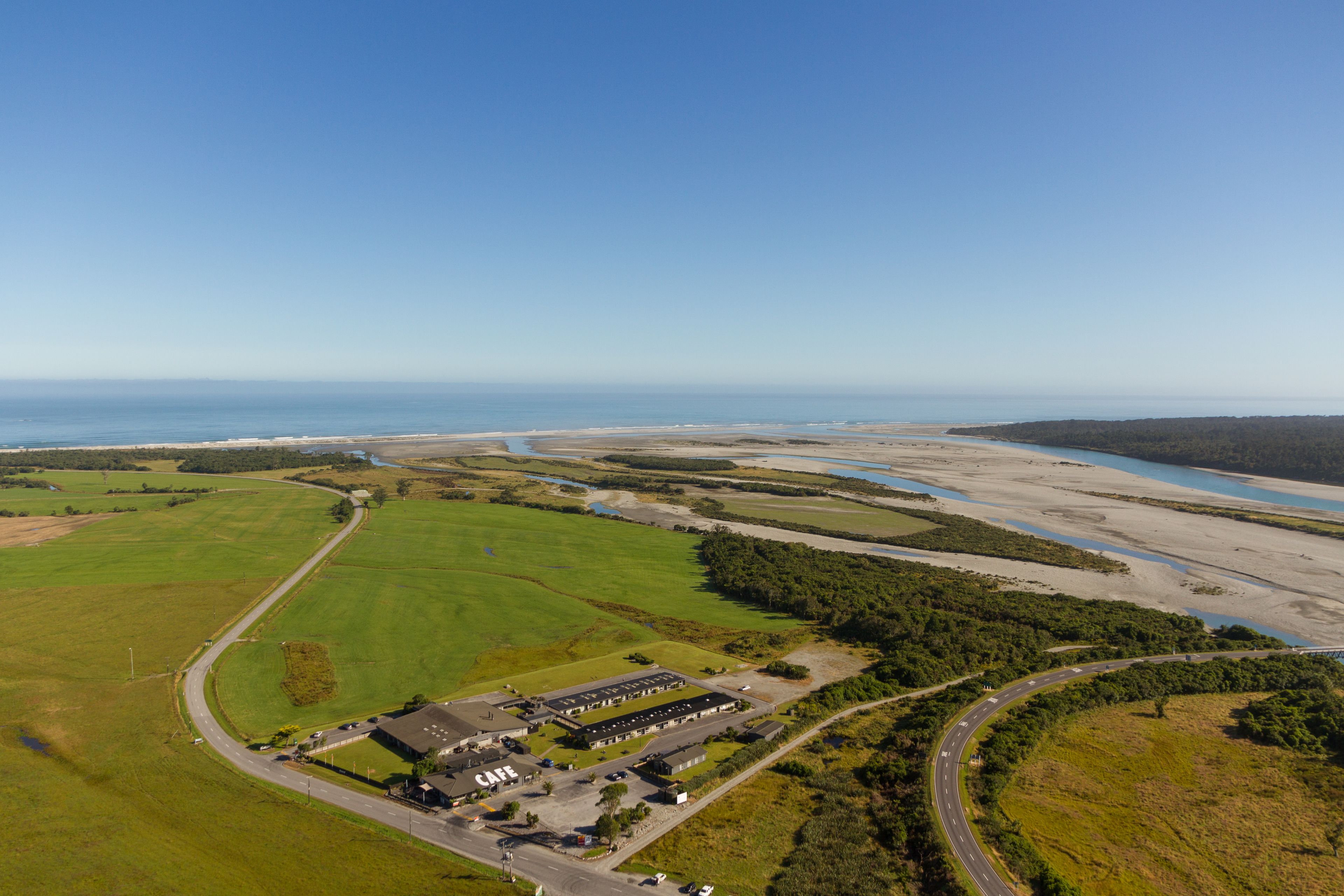Vista aérea de una propiedad con un edificio grande que dice "CAFE", rodeado de campos verdes y una carretera.