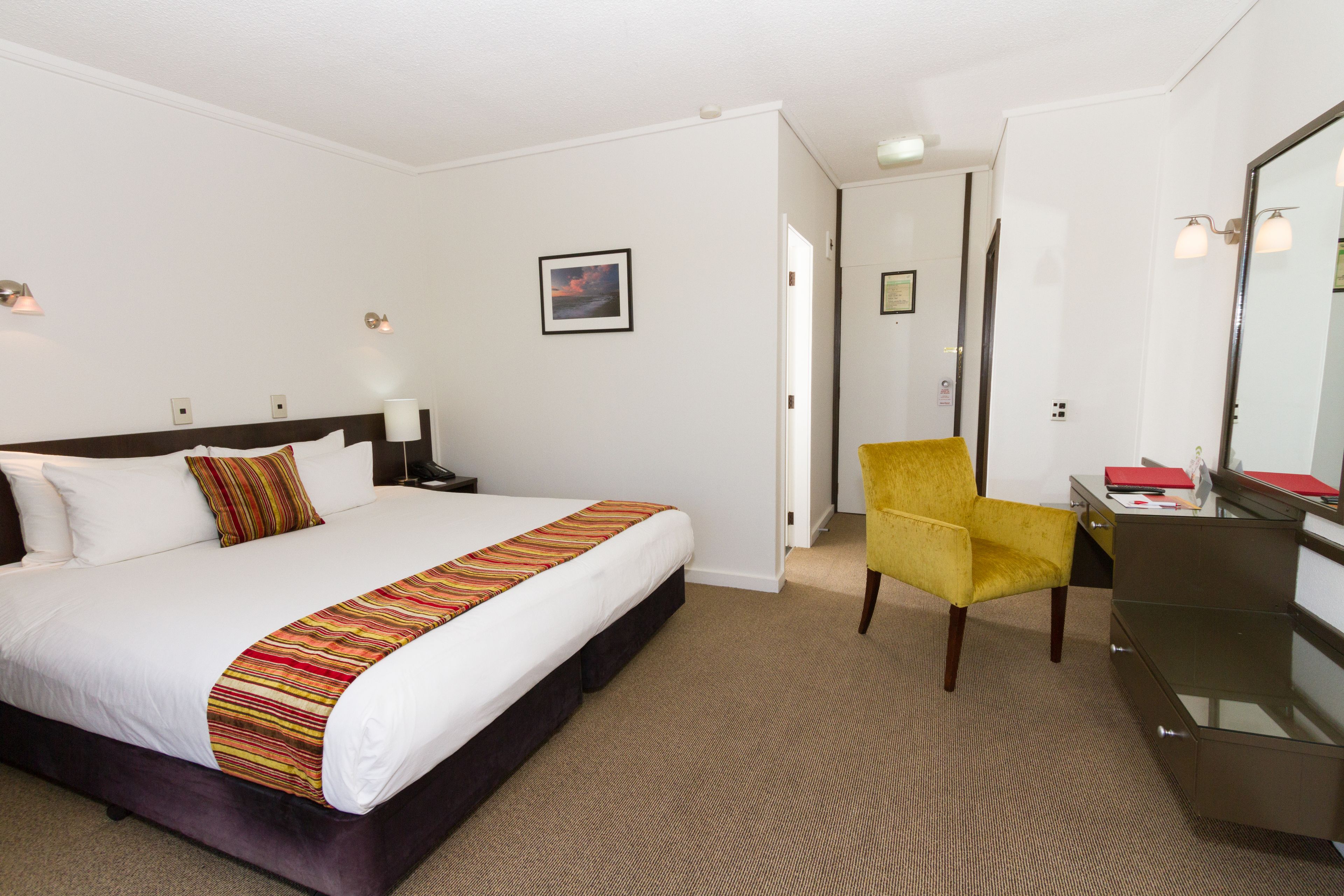 Ein Hotelzimmer mit einem großen Bett, einem Schreibtisch mit Stuhl, einem Spiegel und einer Tür zu einem anderen Raum.