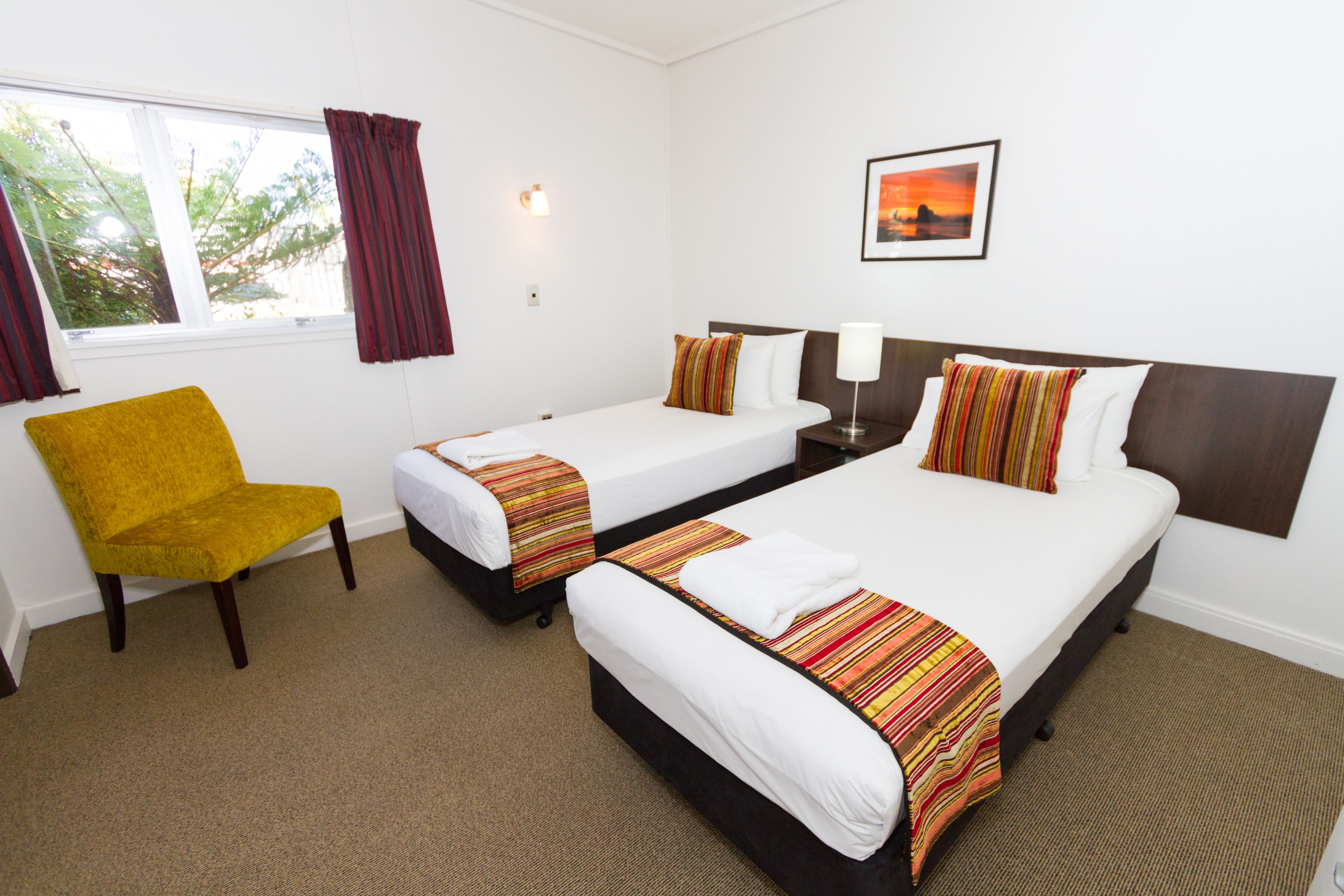 A hotel room with two single beds, a yellow armchair, a nightstand with a lamp, and a window with curtains.
