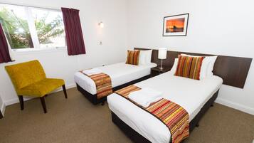 A hotel room with two single beds, a yellow armchair, a nightstand with a lamp, and a window with curtains.