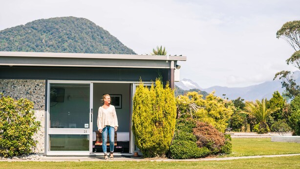 Une maison moderne avec une entrée dotée d’une porte en verre, entourée d’une végétation luxuriante et avec une montagne en toile de fond.