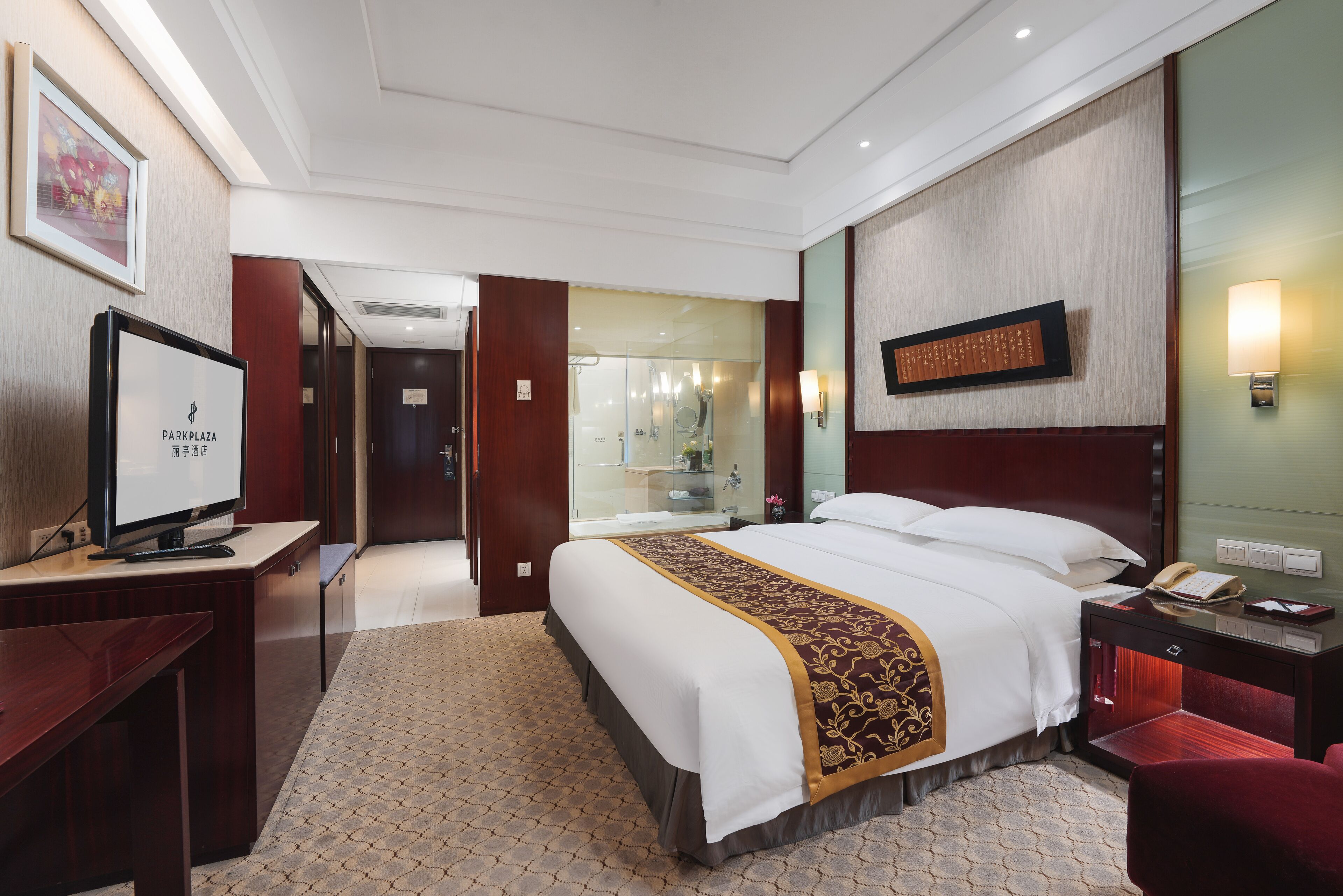 A hotel room with a large bed, a desk with a TV, a mirror, and a bedside table with a lamp.