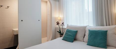 A hotel room with a bed, a window with curtains, a potted plant, and a shower area.