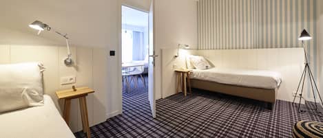 A hotel room with a bed, bedside table, lamp, and a view of a dining area.