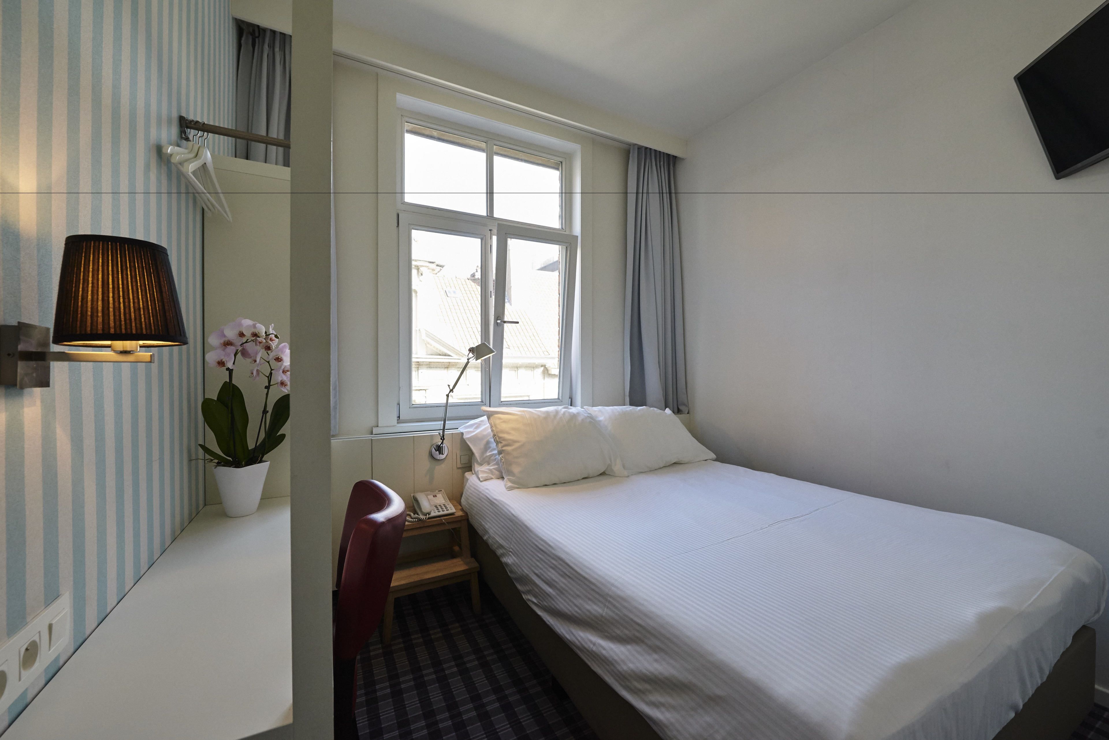 Une chambre d’hôtel avec un lit, un bureau doté d’une lampe, une chaise, une plante en pot et une fenêtre avec des rideaux.