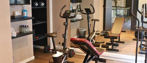 A gym with stationary bikes, a treadmill, and a rowing machine. There are shelves with water bottles and towels.