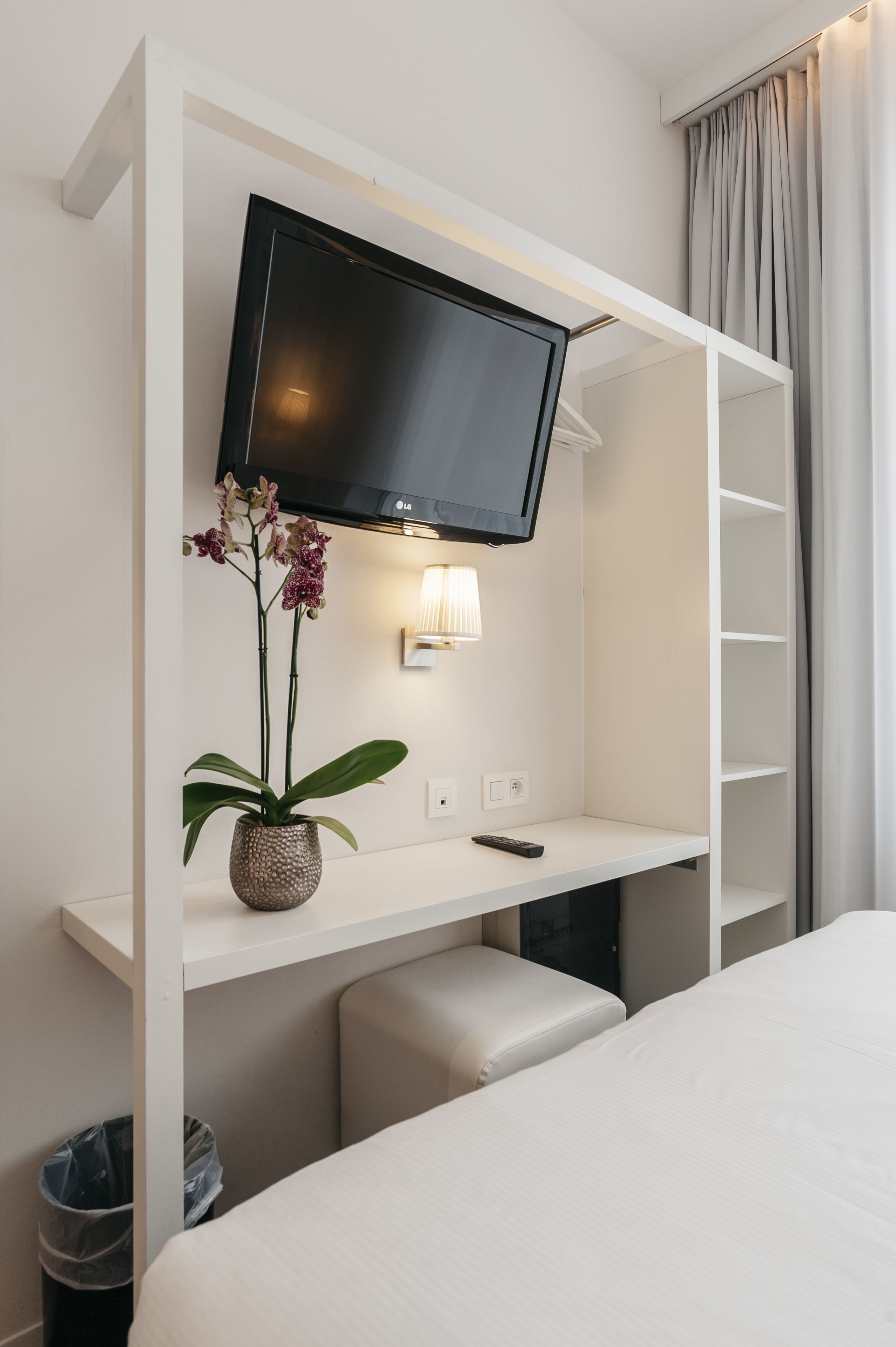 A hotel room with a bed, a TV mounted on a white wall, a small table with a lamp, and a potted plant.