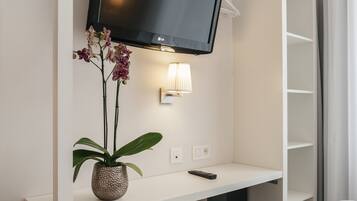 A hotel room with a bed, a TV mounted on a white wall, a small table with a lamp, and a potted plant.