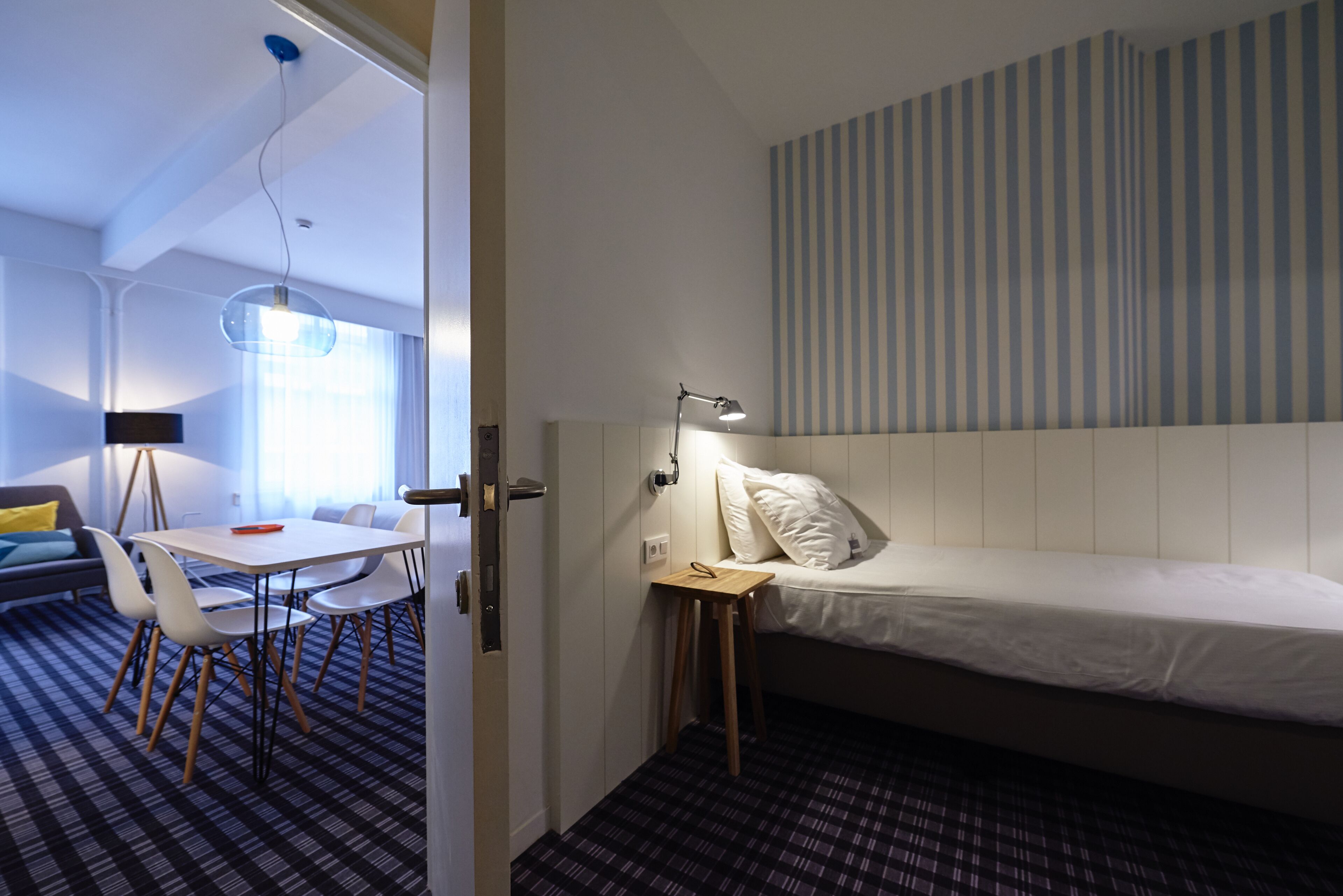 A modern hotel room with a bed, bedside table, lamp, and a dining area with chairs and a table.