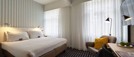 A hotel room with a bed, a desk with a chair, a lamp, and a TV.