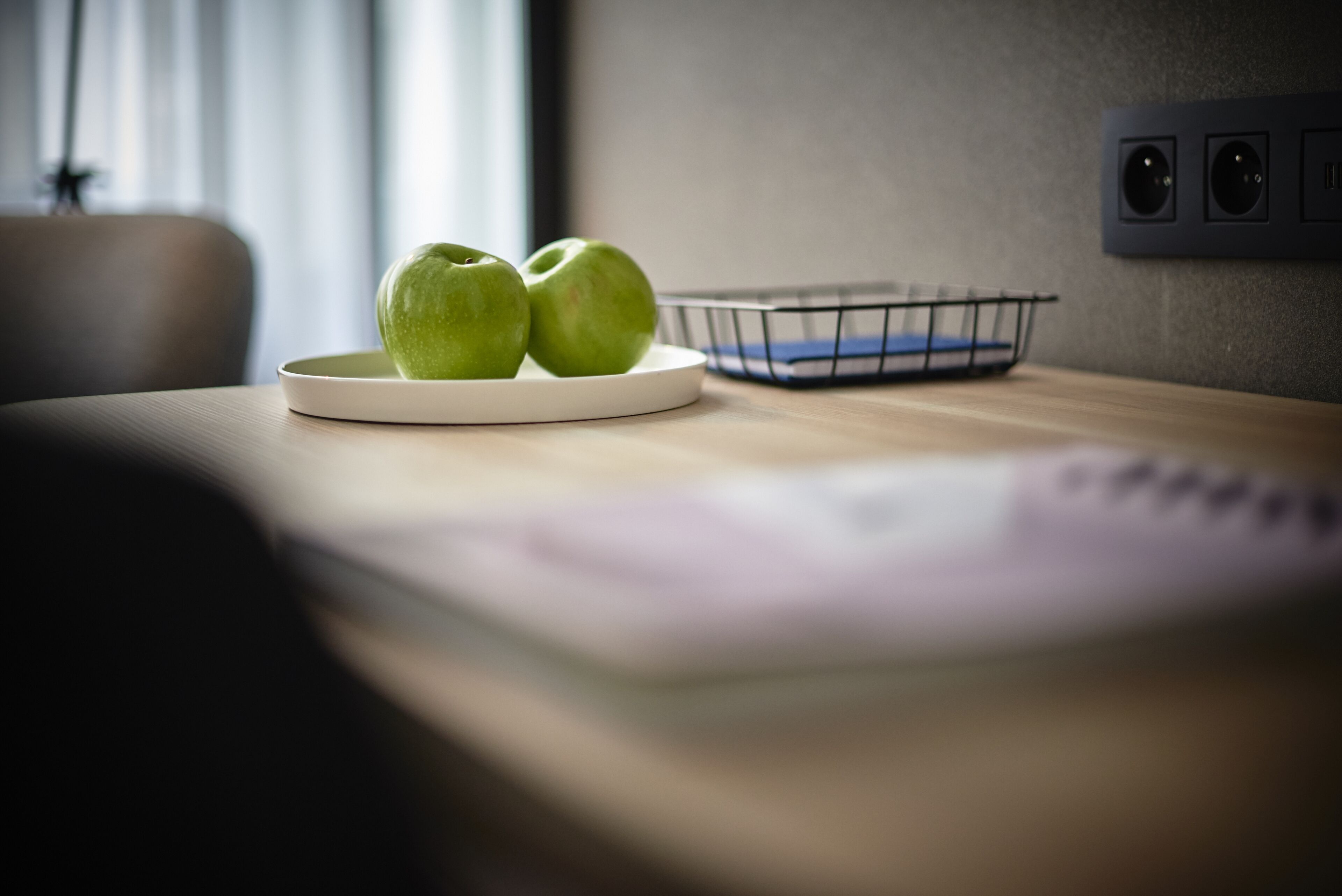 Un plato con dos manzanas verdes sobre una mesa de madera, una cesta de alambre y un enchufe de pared.