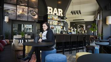 Een bar met een vrouw zittend aan een tafel, een toog met diverse flessen, en een groot verlicht 'BAR' bord.