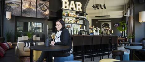 Zona de bar con una mujer sentada en una mesa, una barra con varias botellas y un letrero iluminado grande que dice 'BAR'.