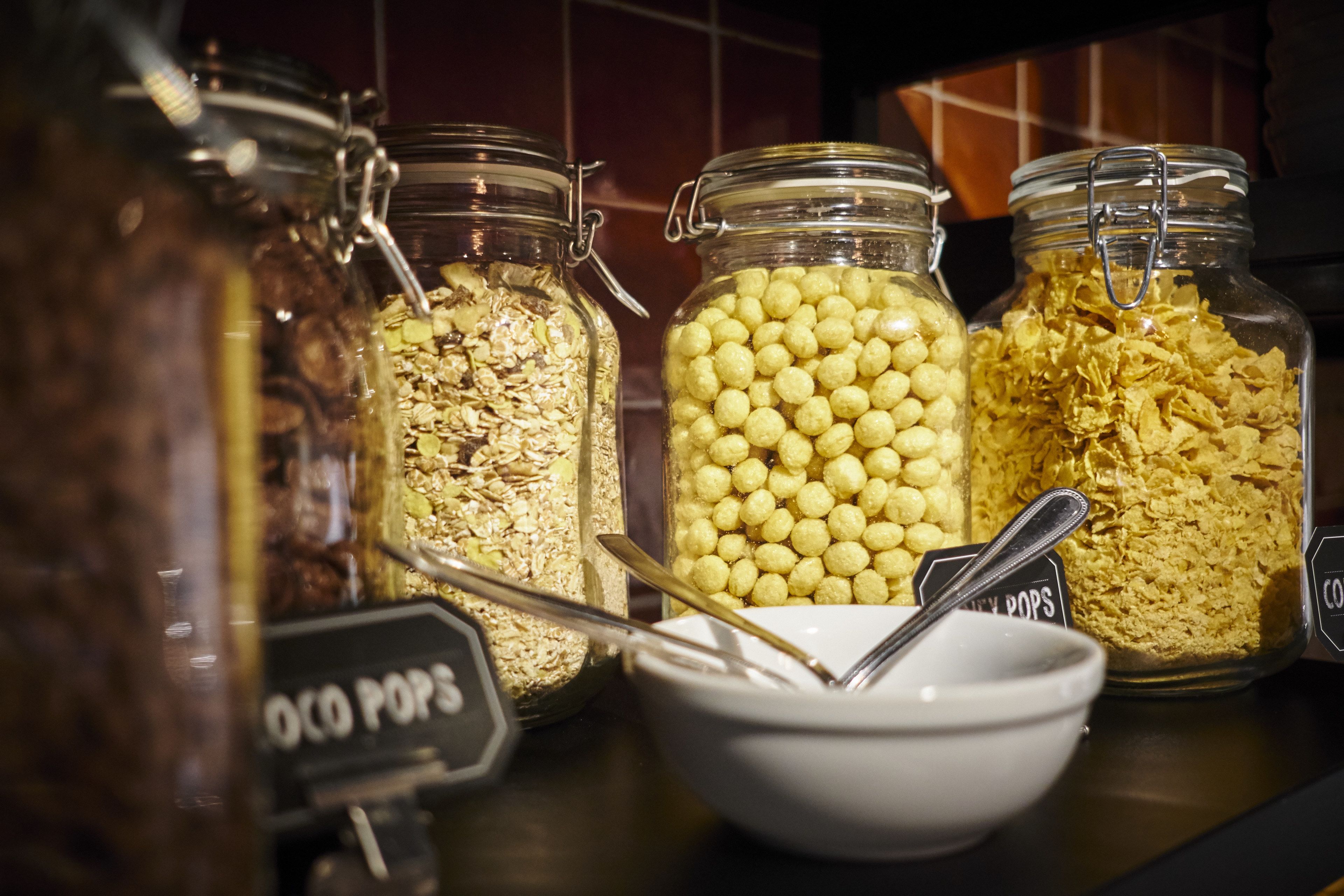 Drei Glasbehälter mit Metallverschlüssen, gefüllt mit verschiedenen Lebensmitteln, eine weiße Schüssel mit einem Löffel und ein Schild mit der Aufschrift 'COCO POPS'.