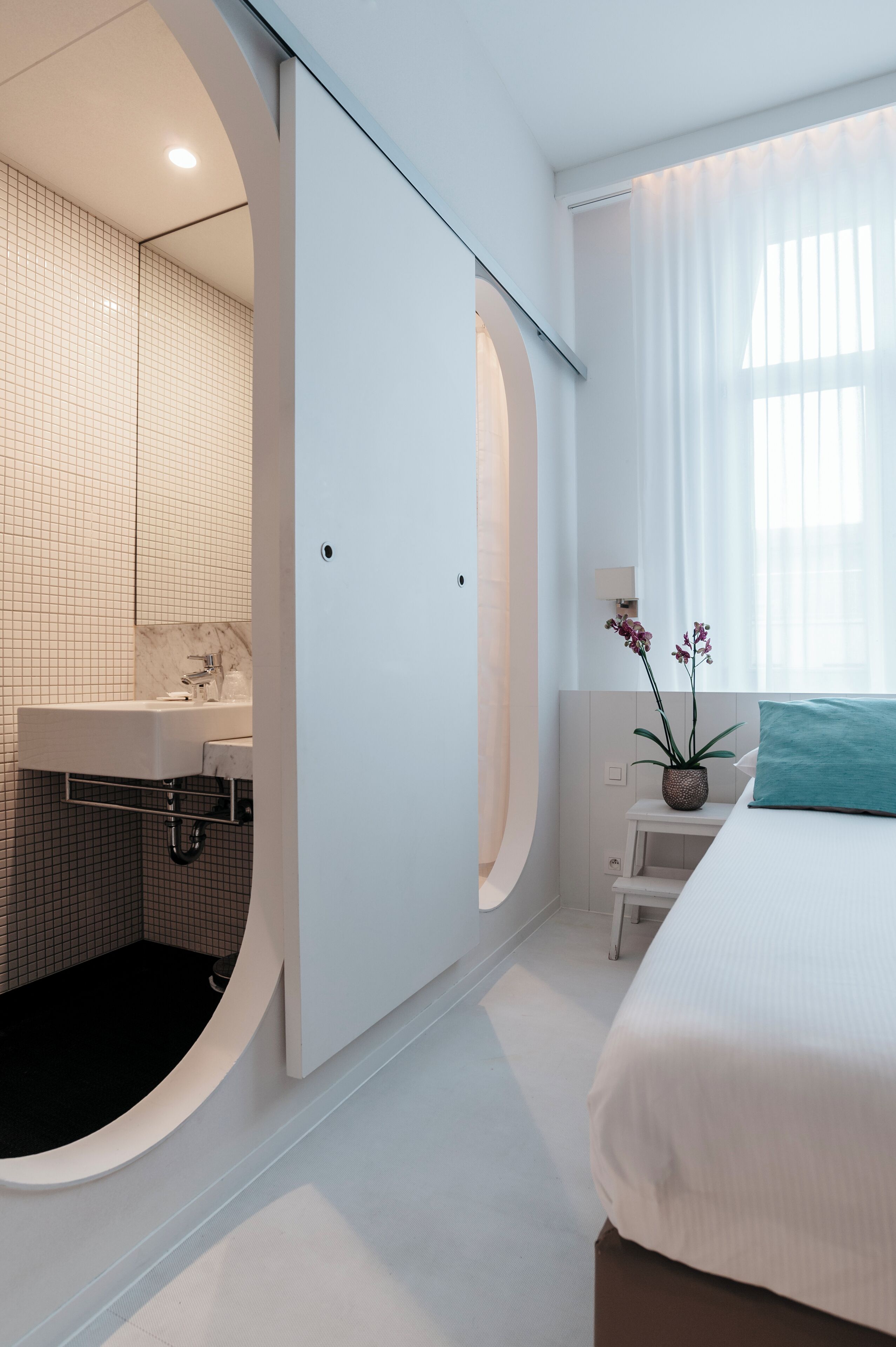 Et moderne badeværelse med en hvid vask, spejl og brusebad. Der er en seng med en turkis pude og et lille bord med en plante.