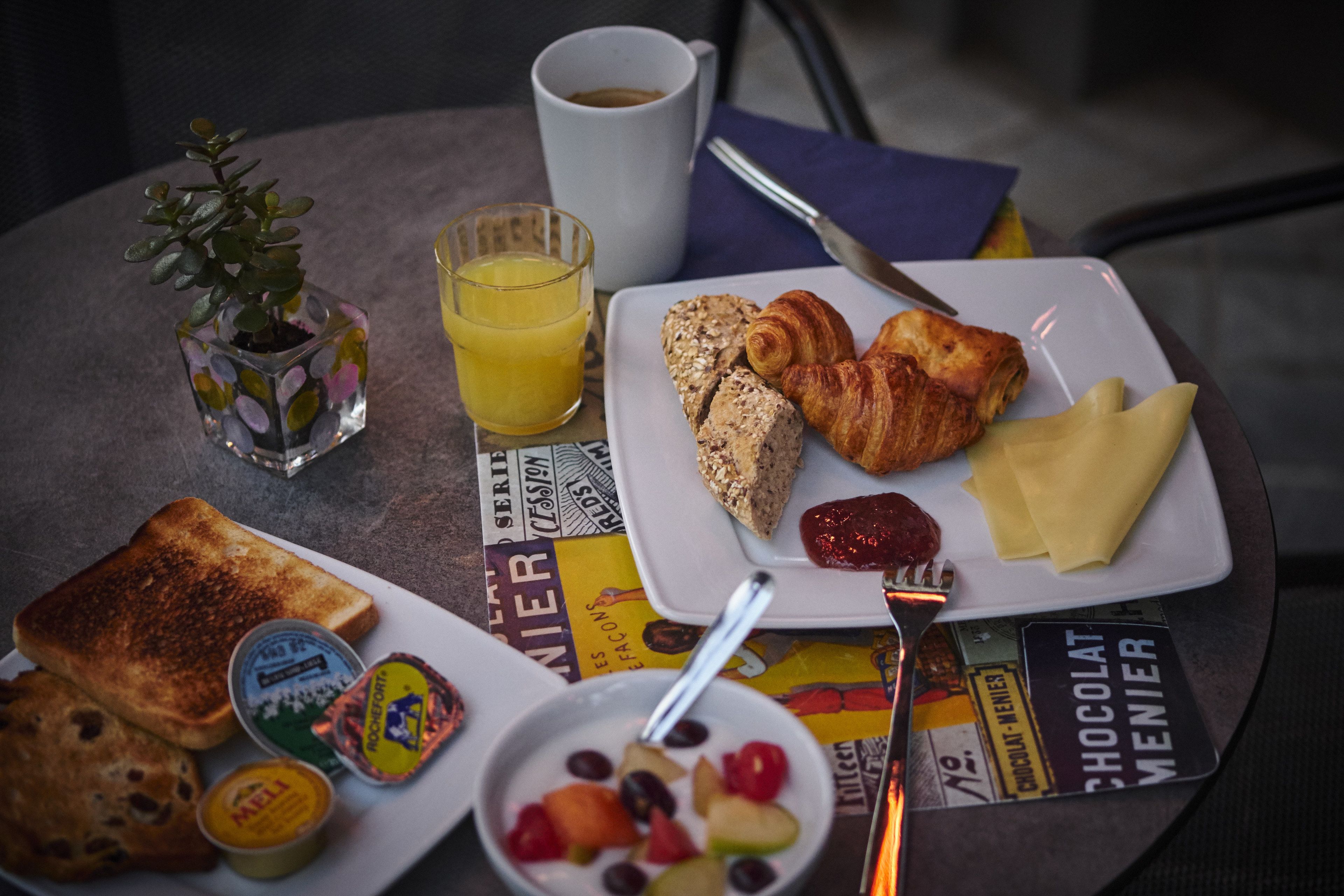 Une table de petit-déjeuner avec des tartines, des croissants, des fruits, un verre de jus d’orange et une tasse de café.