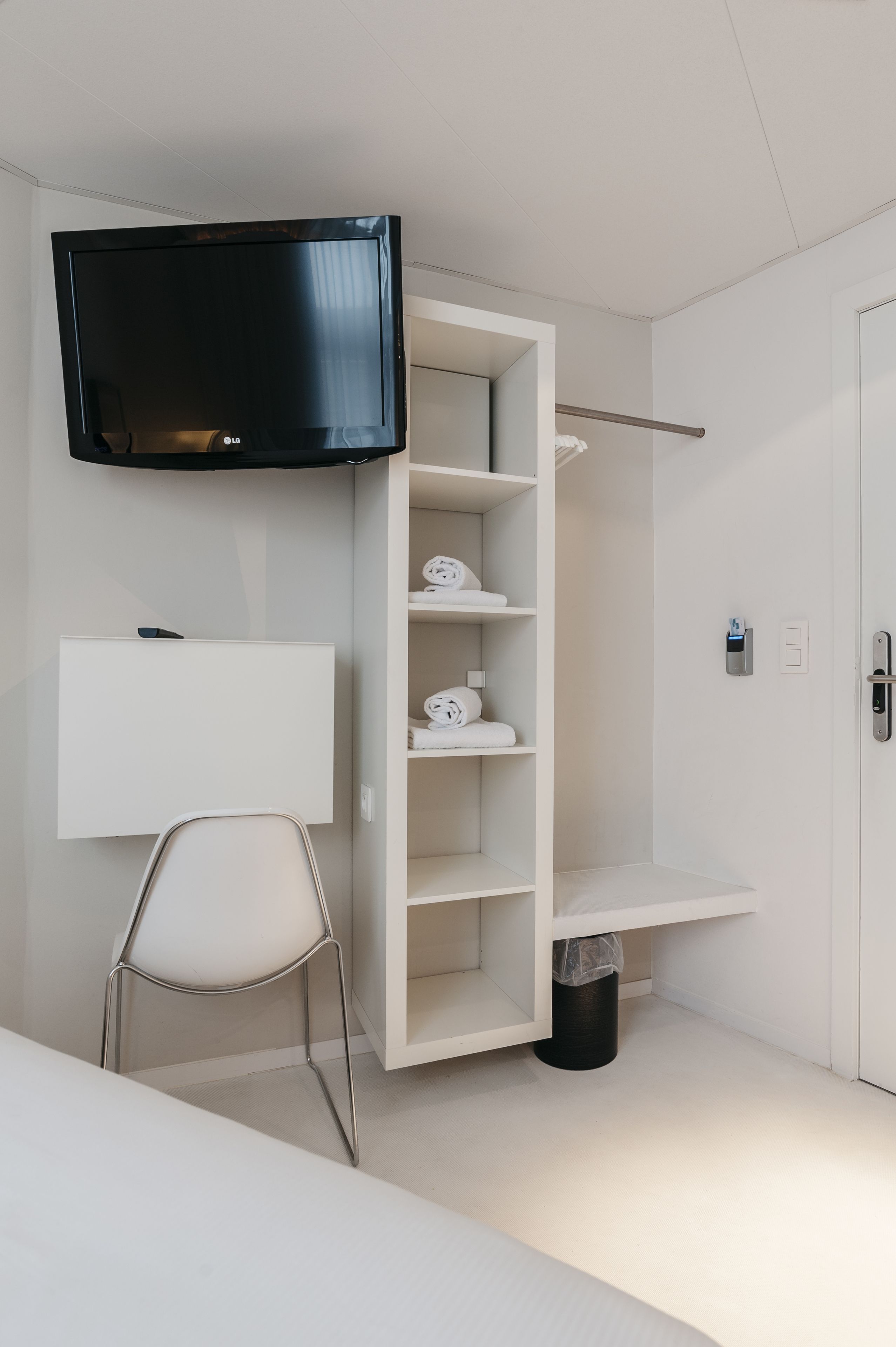 A modern hotel room with a flat-screen TV mounted on the wall, a white chair, a built-in shelving unit, and a door with a keycard reader.