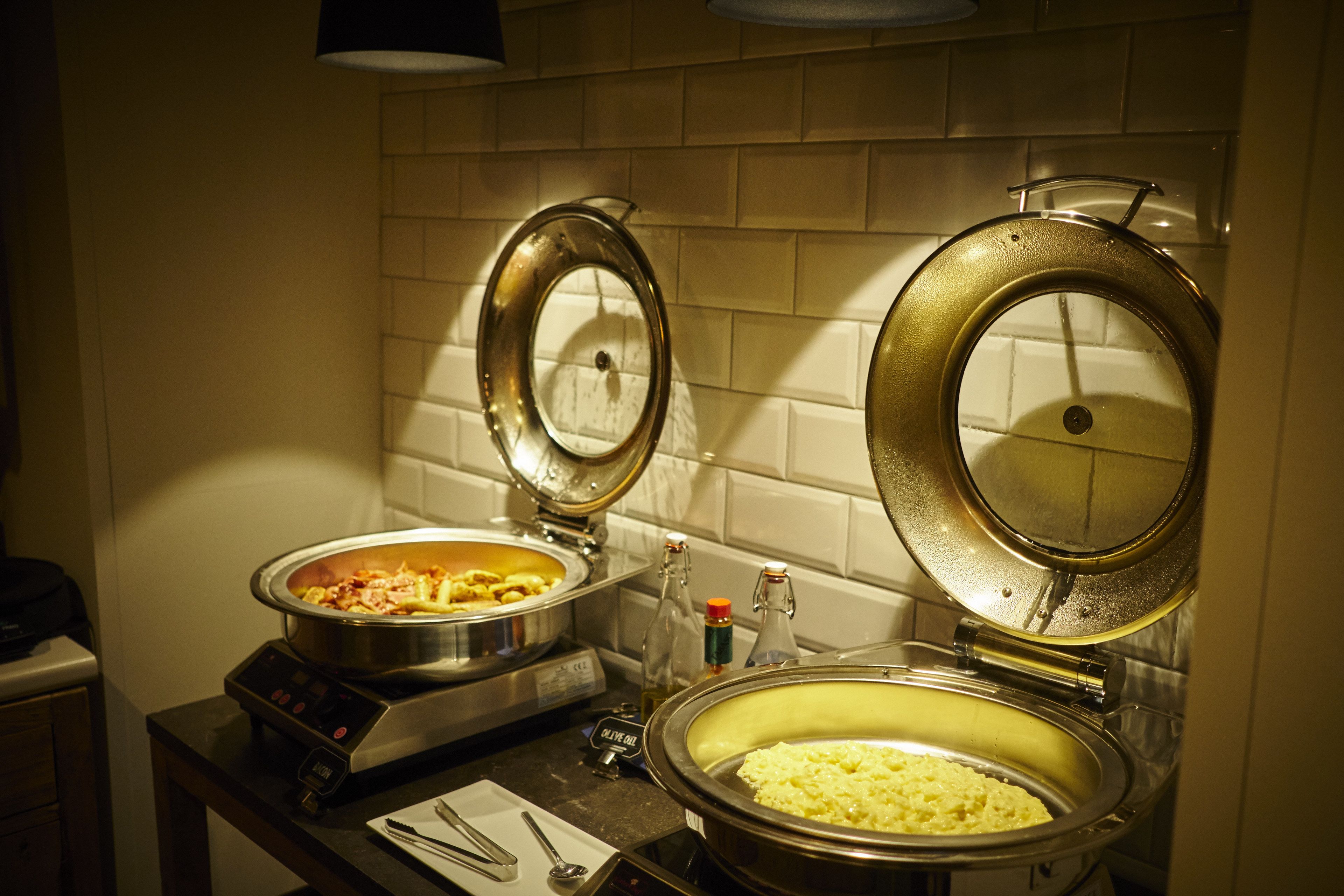 Een buffetopstelling met twee grote schalen, waarvan één gevuld met pasta en de andere met rijst, in een keuken met betegelde wanden en een aanrecht.