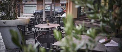 An outdoor cafe with tables and chairs, a large planter, and a view of the street with parked cars and a building in the background.