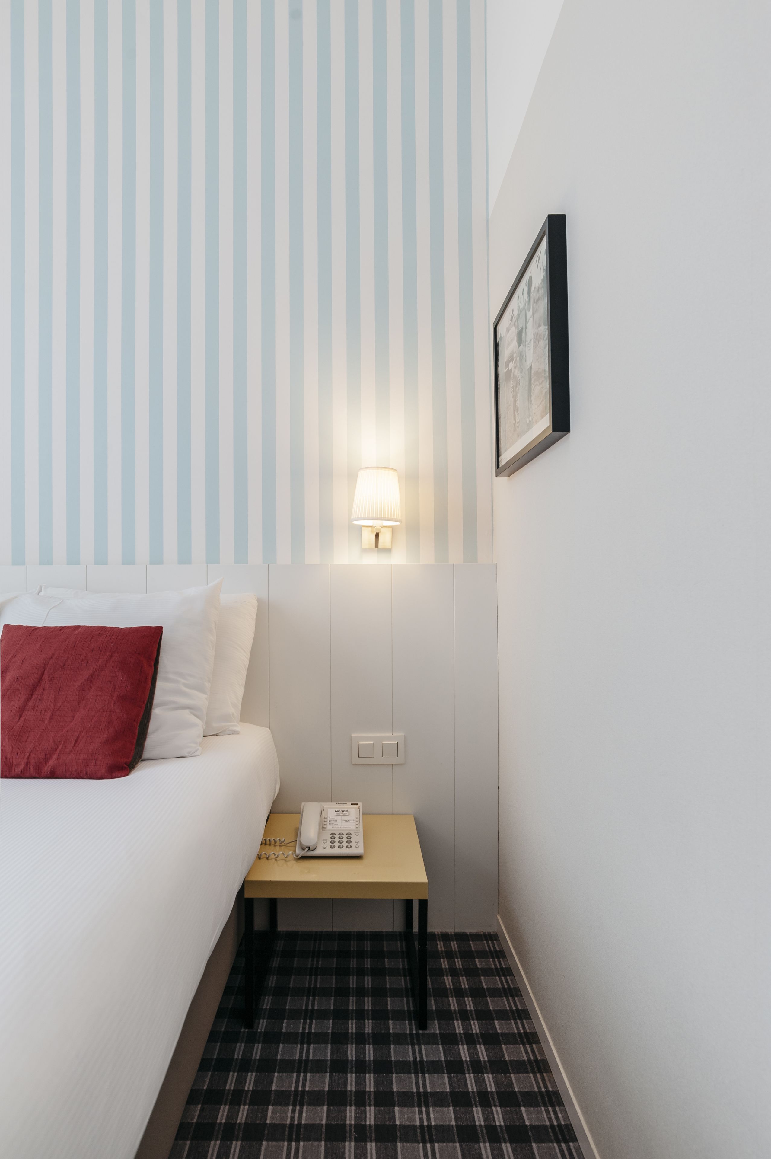 A hotel room with a bed, a bedside table with a lamp, a telephone, and a framed picture on the wall.