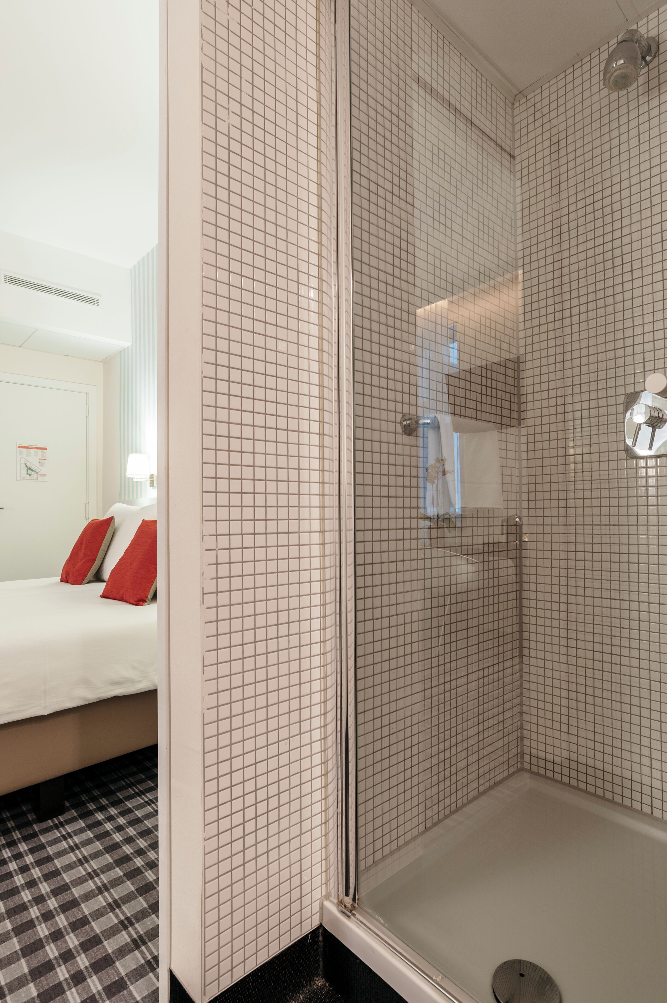 A hotel room with a checkered floor, a bed with red pillows, and a glass-enclosed shower area.