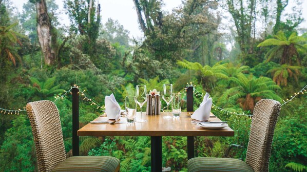 A table set for four with glasses, a bottle, and a flower arrangement, surrounded by chairs, set against a forest backdrop. - Legacy Te Waonui Hotel Franz Josef (Franz Josef Glacier)