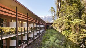 A modern building with a long corridor and multiple balconies, surrounded by lush greenery and mountains in the background. - Legacy Te Waonui Hotel Franz Josef (Franz Josef Glacier)