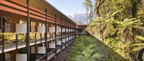 Un edificio moderno con un largo pasillo y varios balcones, rodeado de abundante vegetación y montañas al fondo.