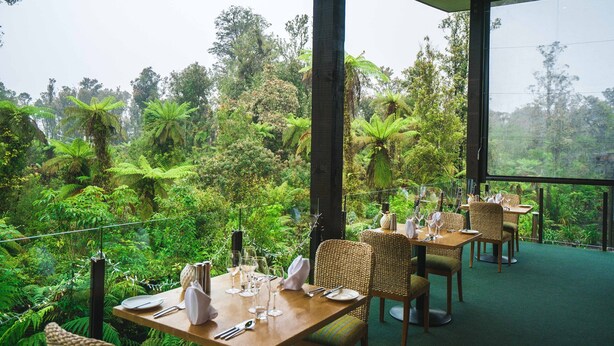An outdoor dining area with tables set for two, surrounded by lush greenery and trees. - Legacy Te Waonui Hotel Franz Josef (Franz Josef Glacier)