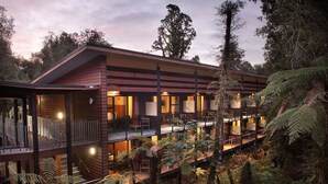 A modern multi-story building with a wooden exterior, multiple balconies, and a lush surrounding forest. - Legacy Te Waonui Hotel Franz Josef (Franz Josef Glacier)